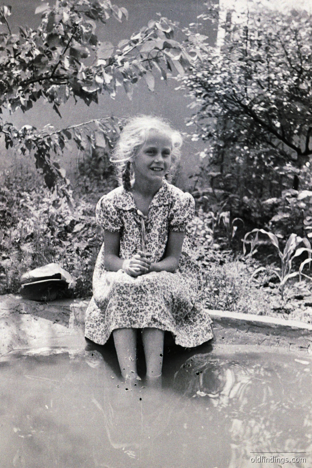 Young girl with braided hair sits in shallow water, wearing a patterned dress. The scene depicts a natural, wooded landscape, likely a pond or marsh. Appears to be a candid portrait, likely taken in the mid-20th century. Image shows signs of age and possible water damage.