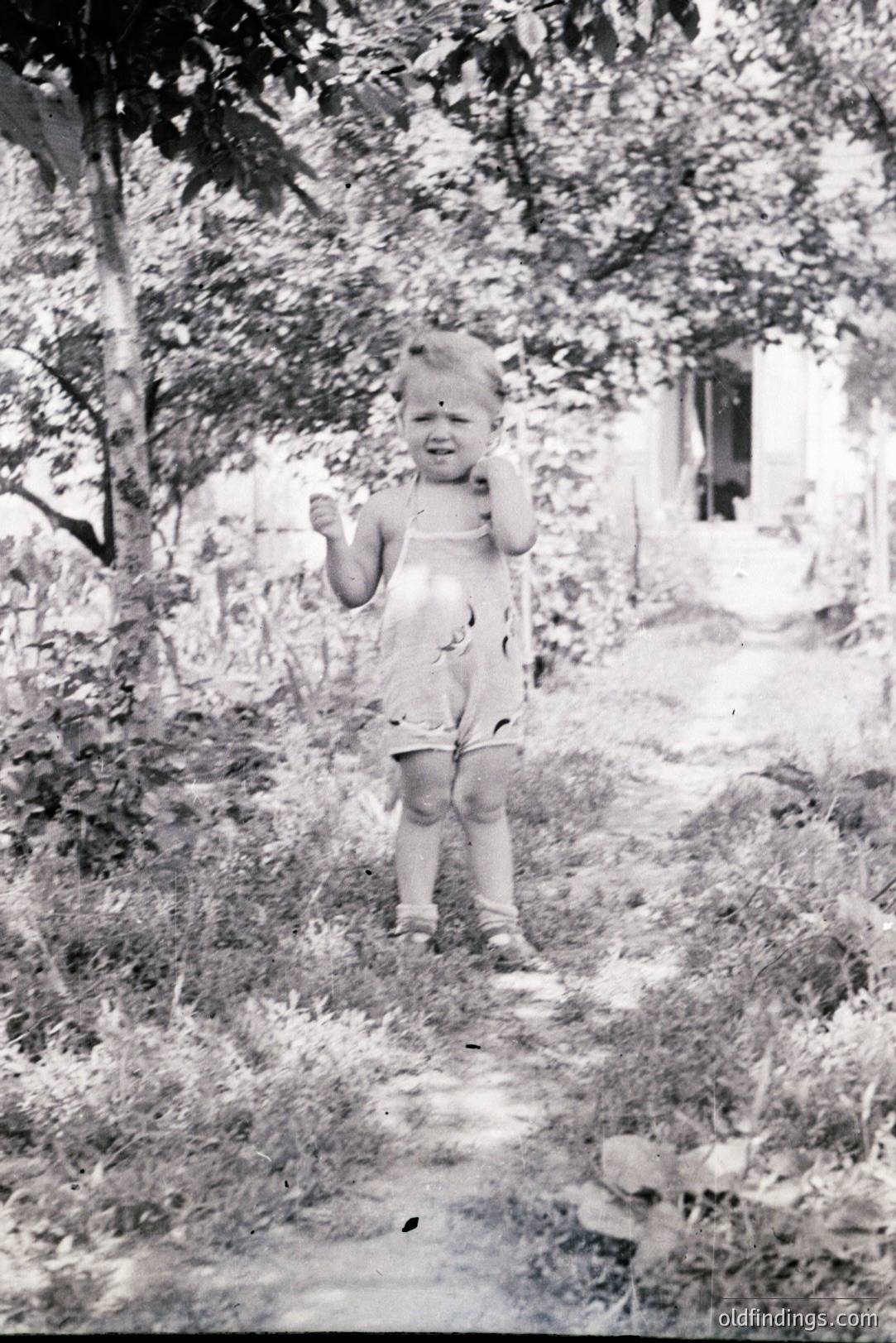 A young child, possibly toddler, stands in a garden path, exhibiting distress – tears and a hand to the face. The child wears a patterned romper and sandals. Surrounding foliage and a glimpse of a building frame the scene. Likely a candid family snapshot, circa 1950s-1970s. Valuable for nostalgic, sentimental, or family history contexts.
