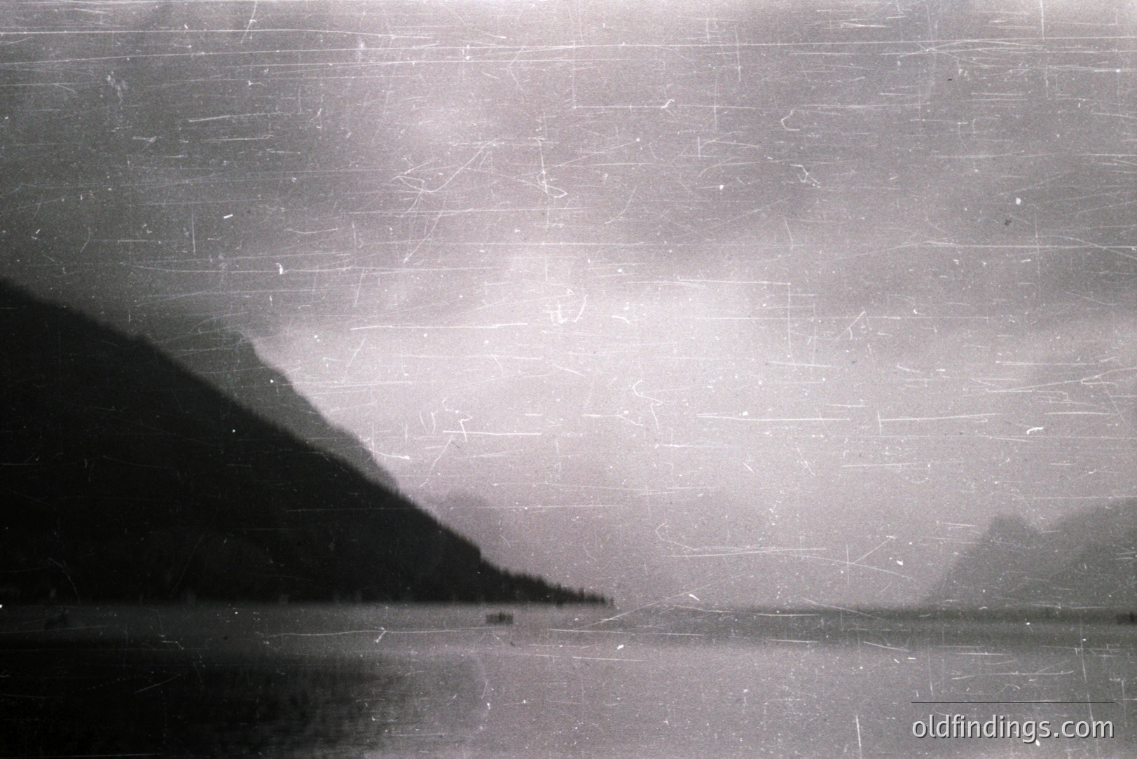 Striking monochrome landscape featuring a dark, forested mountain slope rising from a body of water. Distant, hazy peaks are visible across the water. Image exhibits noticeable film grain and scratches, indicative of age. Likely early-mid 20th century.