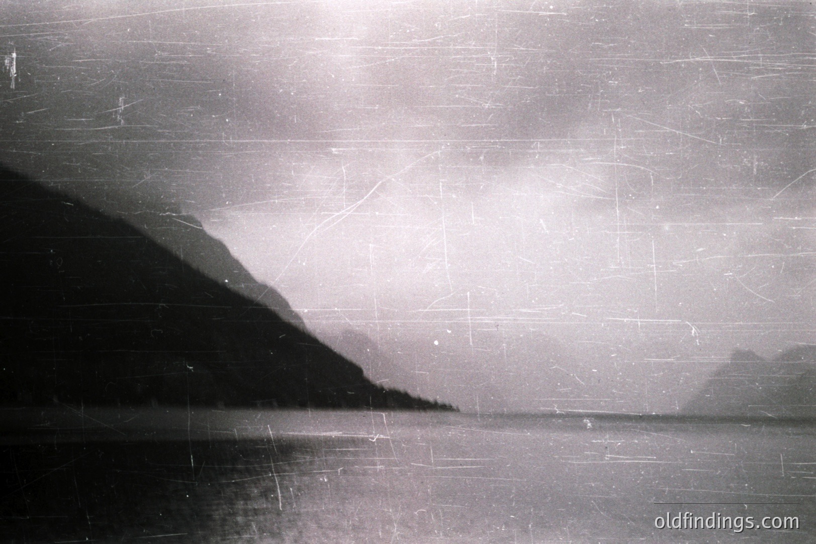 Striking monochromatic landscape showcasing a placid lake reflecting a clouded sky and a dark, steep shoreline. Visible film grain and extensive surface scratches indicate age and potential archival value. Likely taken mid-20th century; location undetermined. Demonstrates stark natural beauty and textures.