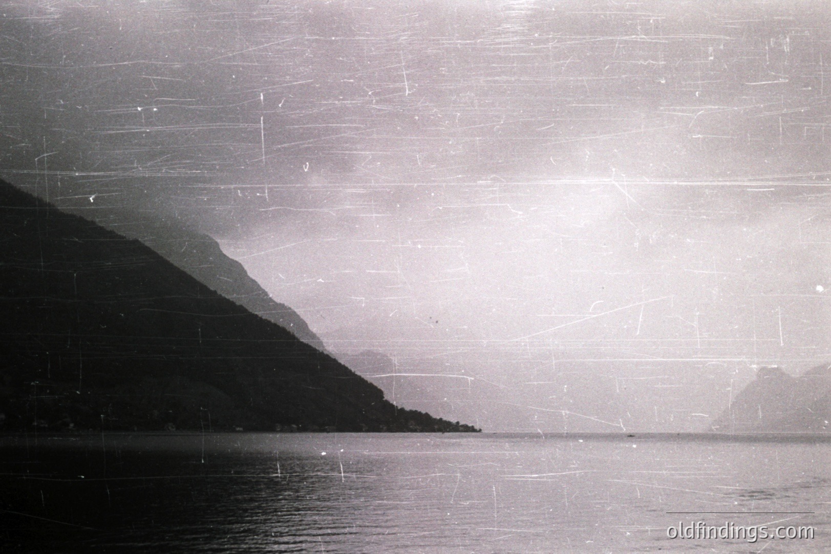Dramatic monochrome seascape. A steep, forested hillside rises from calm water, partially obscured by mist or fog. Visible signs of film grain and surface scratches indicate age. Likely a coastal scene, possibly Scandinavian. Appears to be from mid-20th century stock photography.
