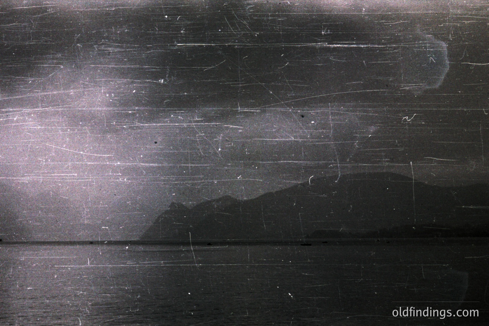Seascape featuring a dark body of water and distant, silhouetted mountains under a cloudy sky. Visible scratches & aging mark the image's surface, suggesting a vintage photograph, likely from the mid-20th century. Evokes a mood of solitude & stillness.