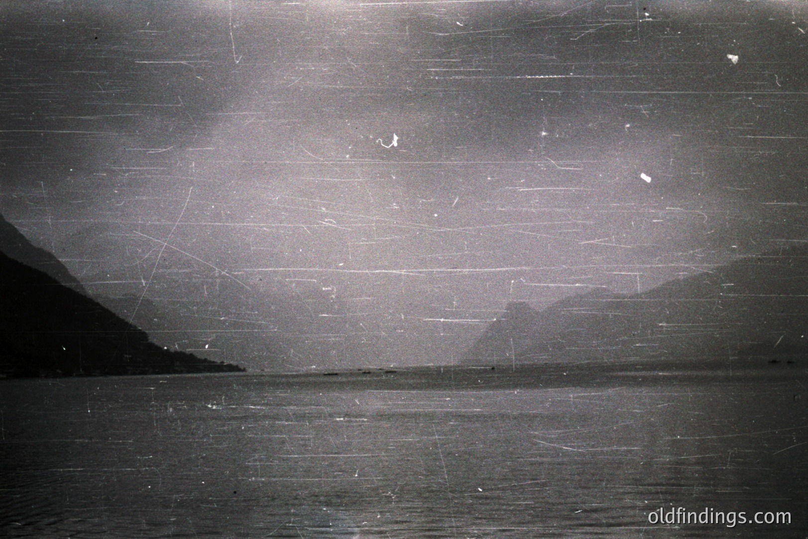 A black and white landscape shows a body of water extending to a distant shore defined by mountains. The image exhibits significant film grain and scratches, typical of older photographic processes. Likely a coastal scene, possibly from the mid-20th century. Offers a vintage aesthetic for design or historical research.
