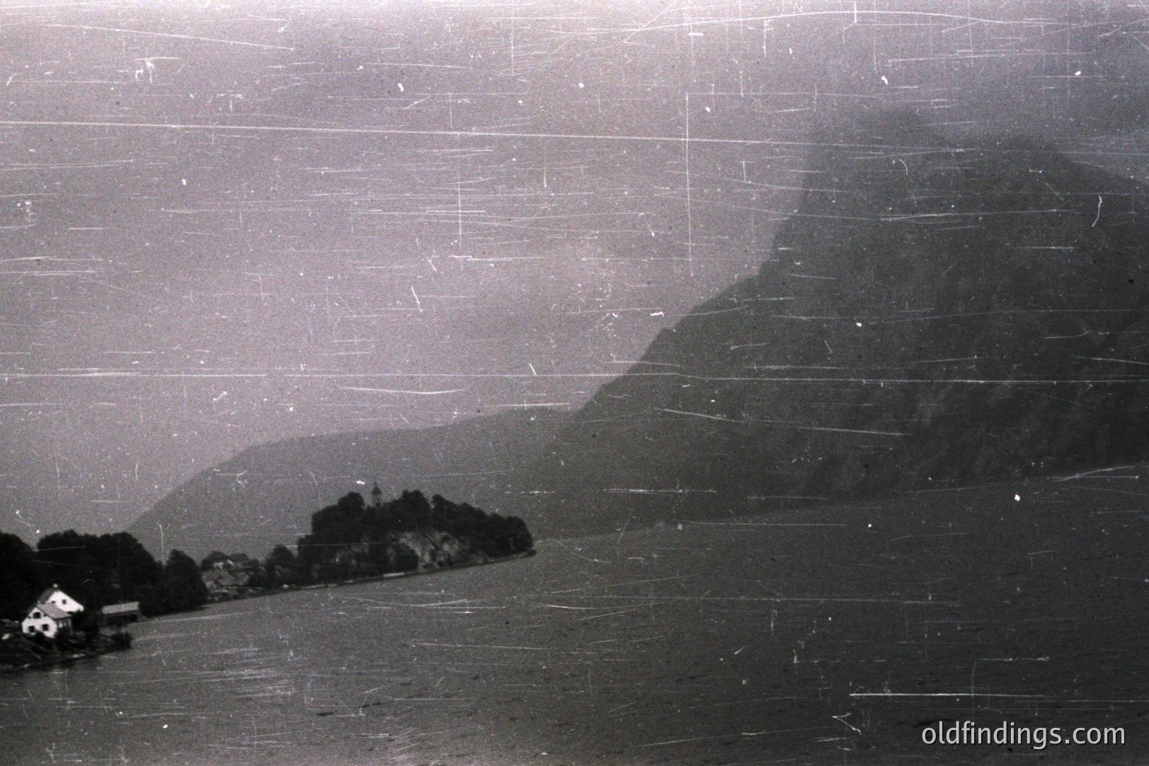 A hazy, grayscale view of a fjord landscape with a large mountain rising on the right. A small, white building sits on the shoreline. Extensive scratches likely from age/handling. Suggests a mid-20th century travel photograph, possibly Scandinavia. Useful for design referencing or historical context.