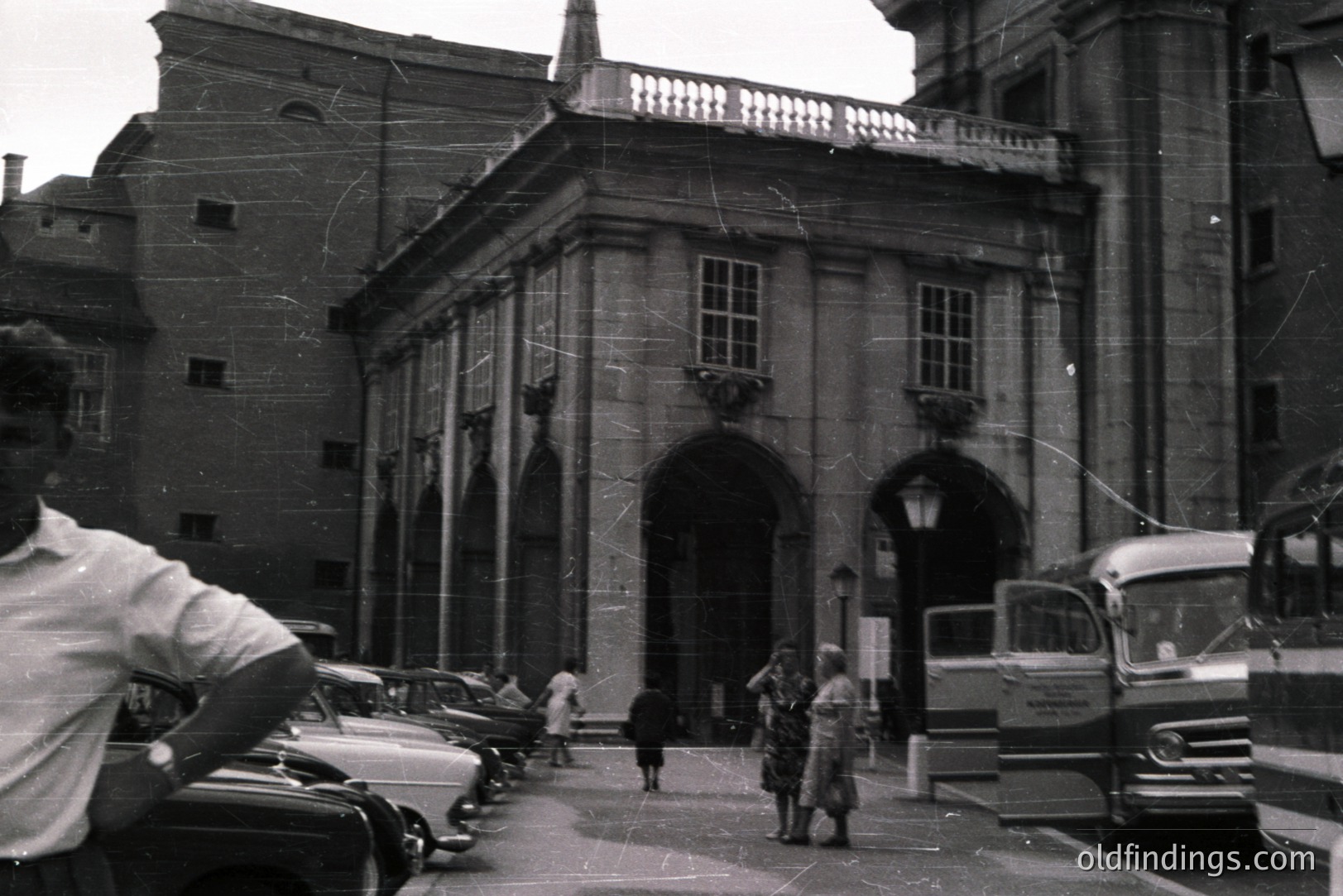 Classic architectural facade with arched portico and balcony, framed by surrounding buildings. Several vintage cars and a bus are parked along the street. Figures are visible in the courtyard and walking by. Likely 1960s, European location. Interesting streetscape & urban design reference.