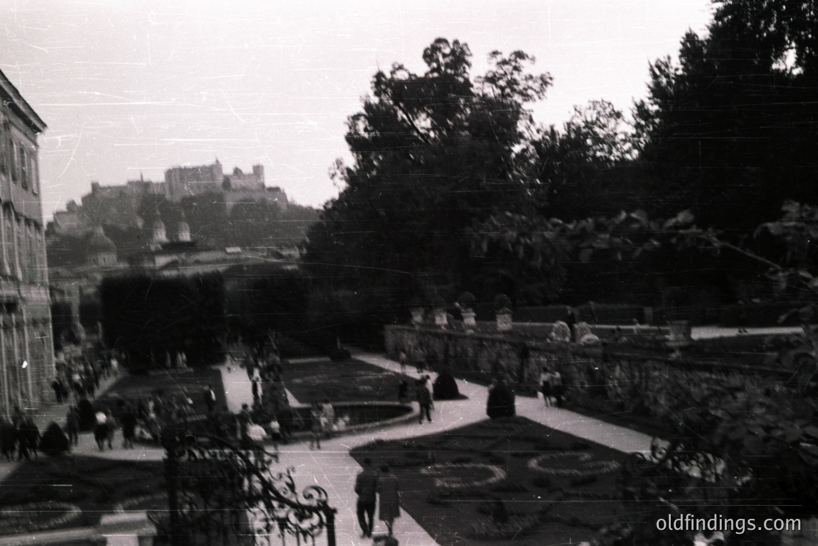 Formal garden featuring manicured hedges, patterned flowerbeds, and stone pathways. A distant castle silhouette dominates the skyline. People are strolling through the gardens. Likely a European estate, circa 1960s-1970s, judging by photographic style. Ideal for landscape or architectural reference.
