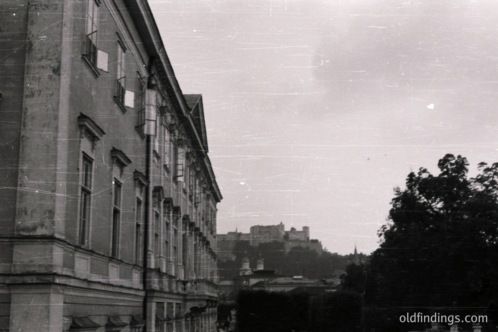 Striking black and white image captures a grand building facade with symmetrical windows and ornate detailing. A distant castle sits atop a hill. Architectural style suggests late 19th/early 20th century, possibly Central Europe. Noticeable film grain and surface scratches.