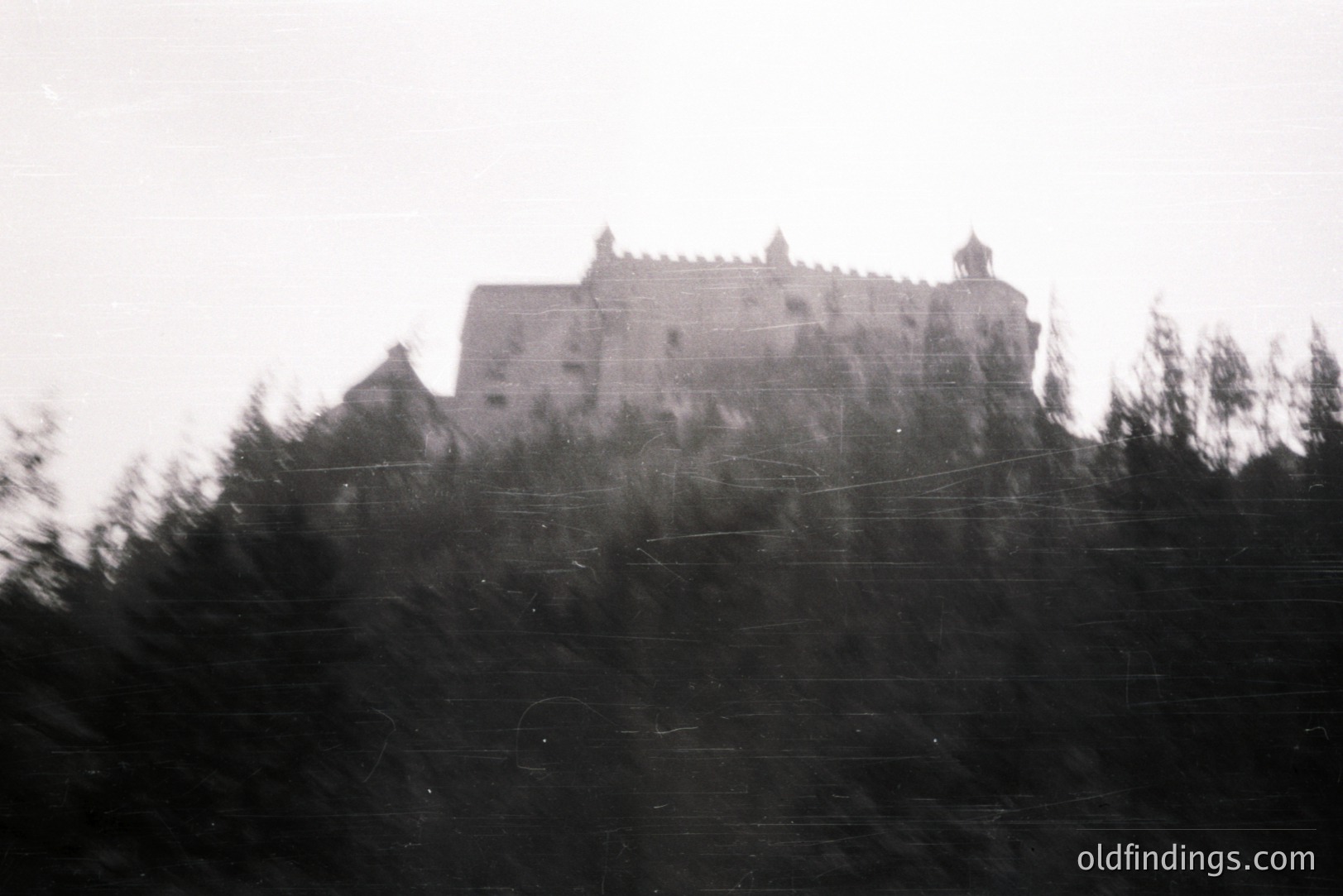 Monochrome photo captures a castle atop a hill, partially obscured by trees. Architectural details show a complex, multi-tiered structure with what appears to be a central tower. Visible signs of age and wear. Likely early to mid-20th century, possibly travel photography.