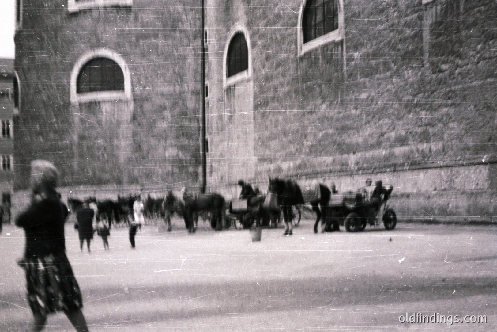 A bustling plaza with a stone building façade featuring arched windows. A horse-drawn cart and several horses are central. A figure appears blurred in motion toward the left. Likely early 20th century, potentially European. The scene suggests a historical market or transportation hub. Appears as a candid moment captured in a documentary style.