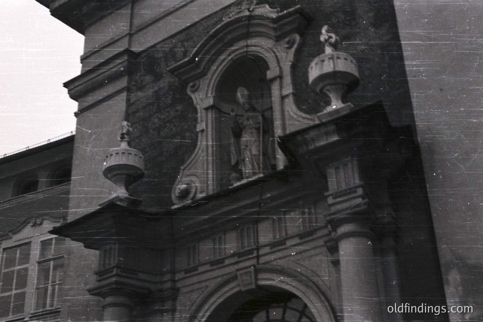 Ornate stone archway & statuary, likely part of a European ecclesiastical building. Detailed carvings exemplify Baroque or Rococo architectural style. Shows signs of weathering. The building's facade reveals multiple windows above. Appears to be a historical photograph, potentially from the early 20th century.