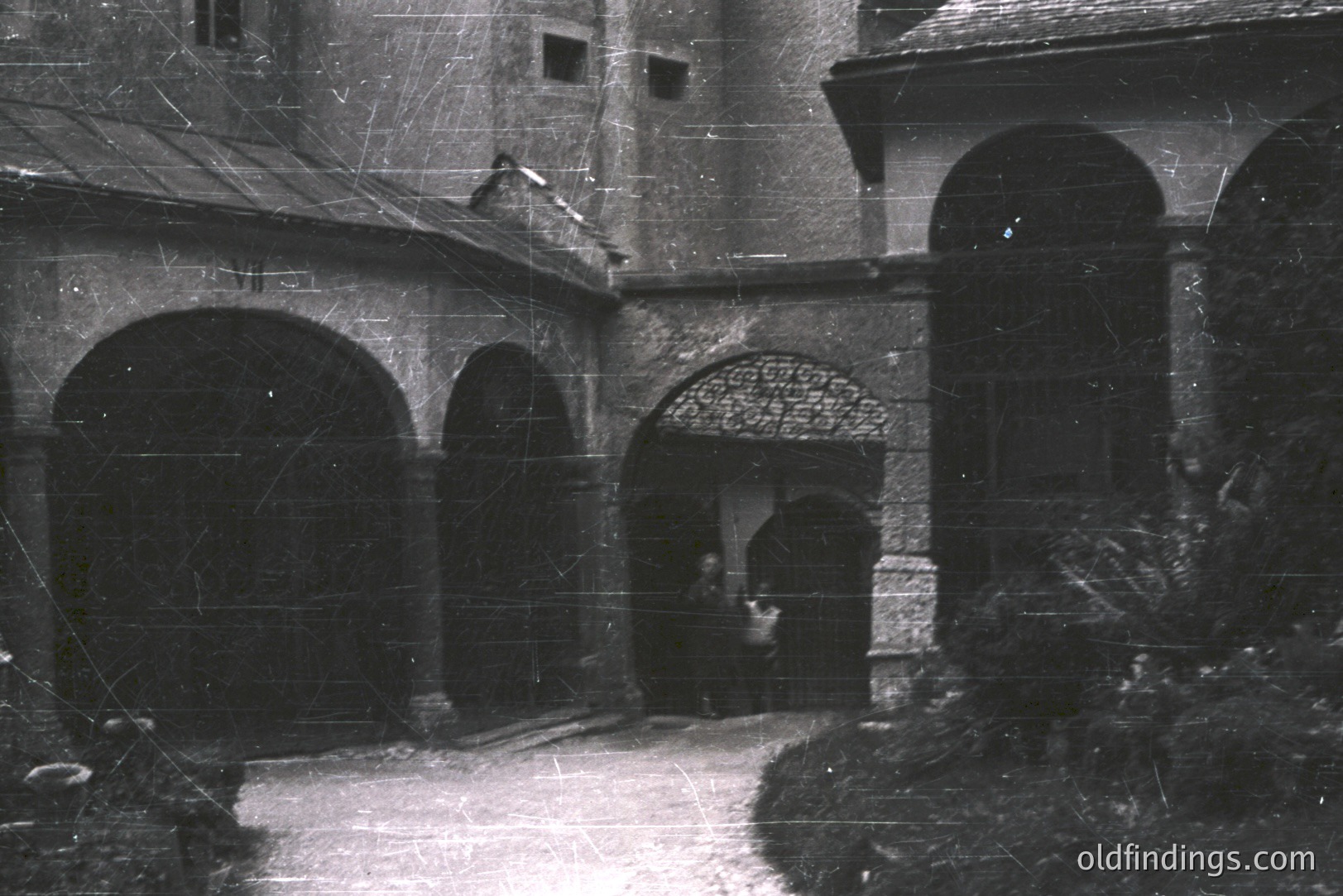 Series of arched passageways form a courtyard with stone paving, leading to a distant arched entrance. A person stands near the far entrance. Likely a European estate or castle. B&W photography suggests a vintage aesthetic, possibly 1930s-1960s. Strong architectural detail.
