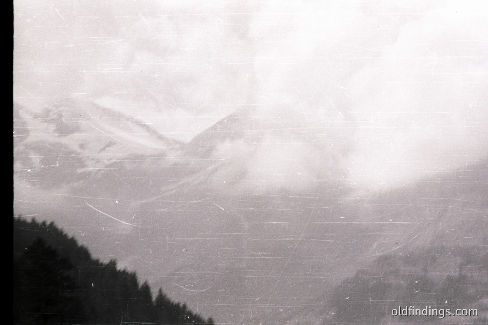 A black and white landscape depicts snow-covered alpine peaks shrouded in mist. Dense evergreen forest occupies the lower frame. Visible scratches and dust artifacts suggest age and film degradation. Likely a scenic view, possibly from the 1960s or 70s.
