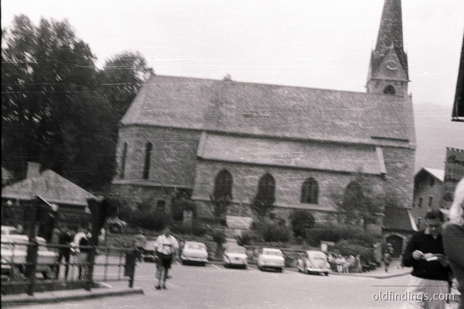 A stone church with a tall steeple dominates this view, situated on a slight rise overlooking a street lined with vintage automobiles. Pedestrians are present. Appears to be a rural European setting, likely 1960s. Potential stock photo for architectural, travel, or historical uses.