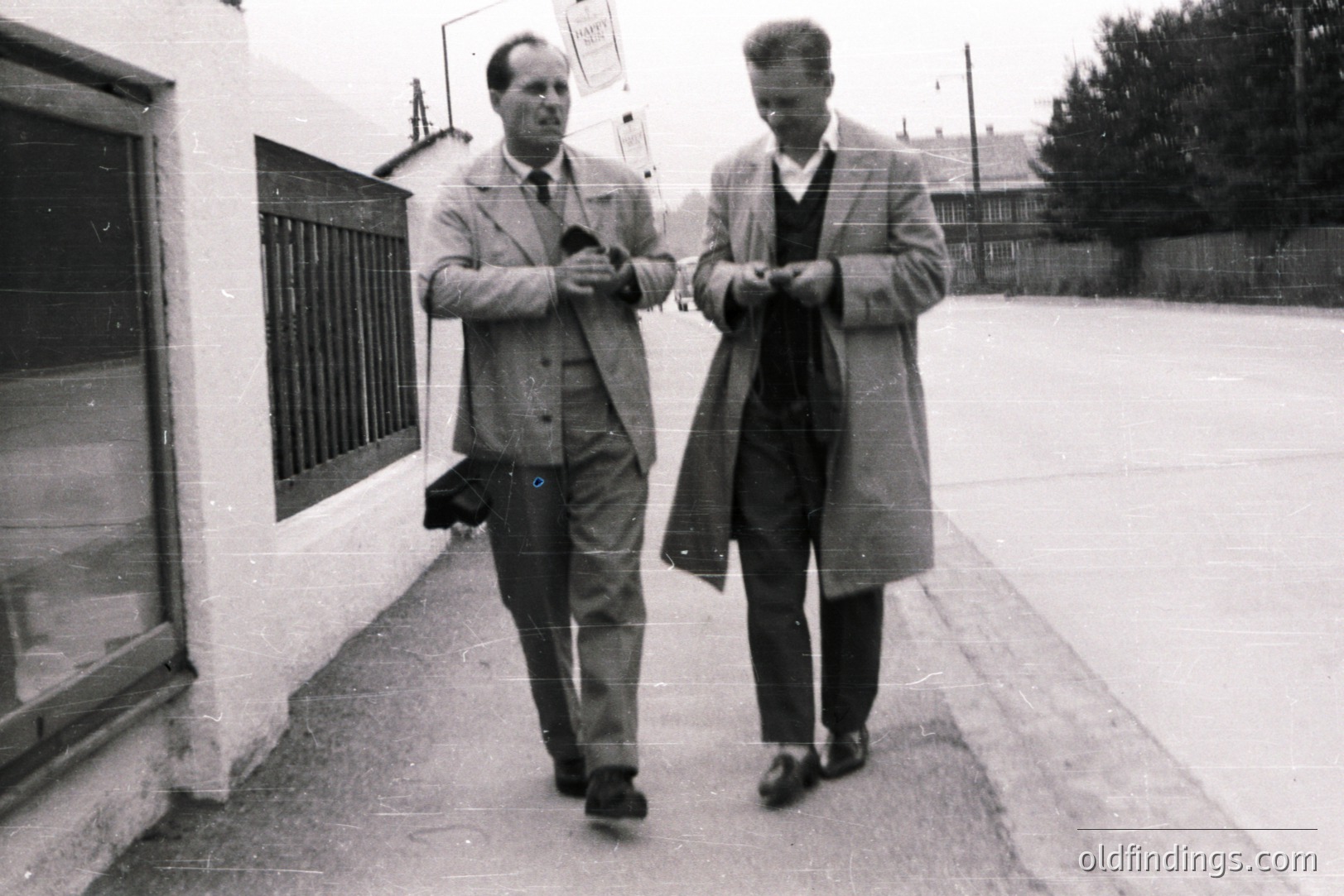 Two men in overcoats walk along a sidewalk, appearing deep in conversation. Architectural details suggest a residential area, possibly suburban. The image displays a grainy texture, indicative of a vintage photograph, likely 1960s or 70s. Commercial value for period design or stock photography.