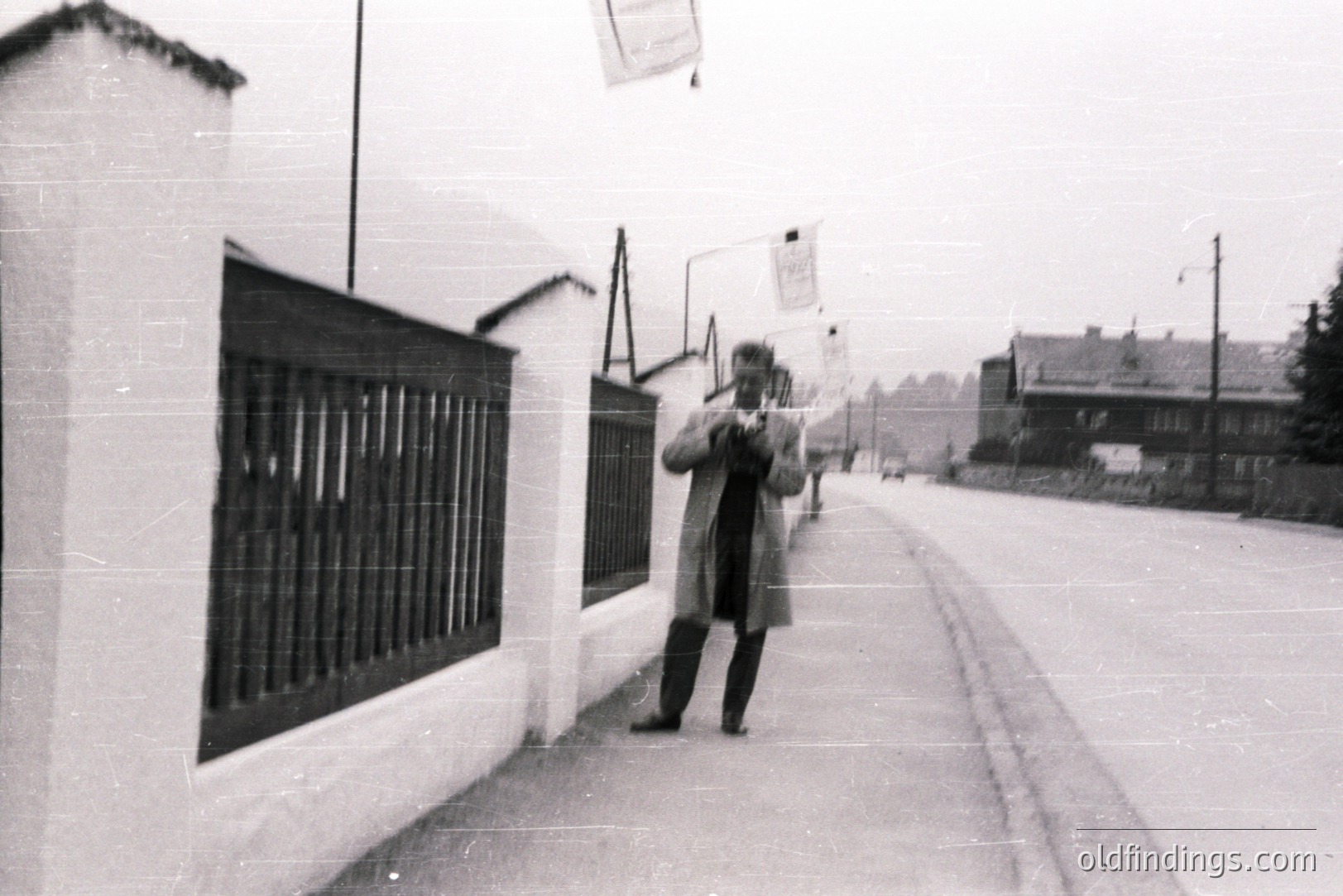 B&W street scene; a man in a long coat stands photographing with a vintage camera. Whitewashed wall & gate dominate left. Sparse, overcast sky & residential buildings in background. Likely mid-20th century. Architectural style suggests central or eastern European influence. Appears to be a documentary or personal snapshot.