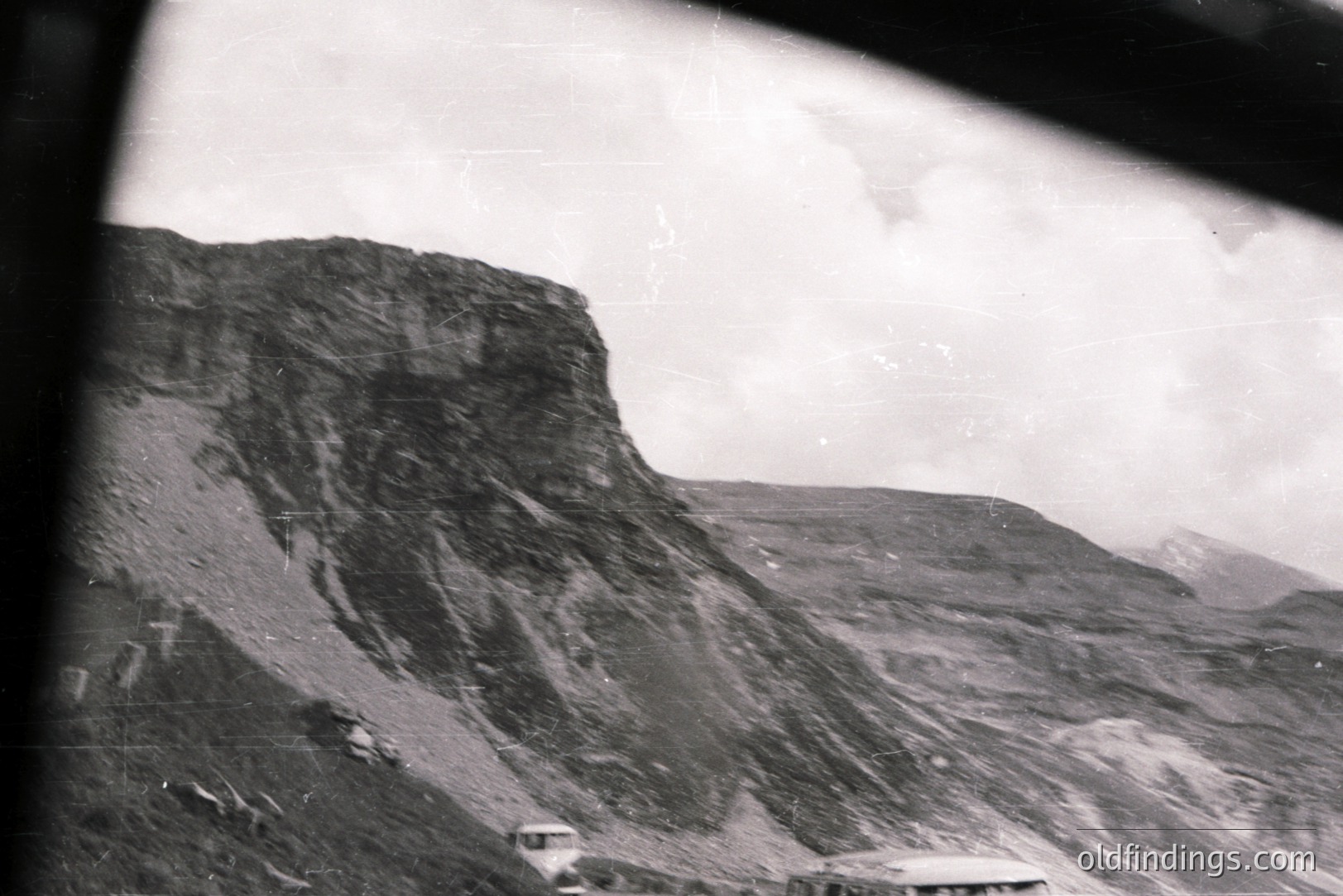 Striking alpine landscape with rugged, layered rock formations and sparse vegetation. A distant structure suggests a mountain lodge or observation point. Visible film grain & partial frame edge indicate vintage photographic techniques. Likely mid-20th century.