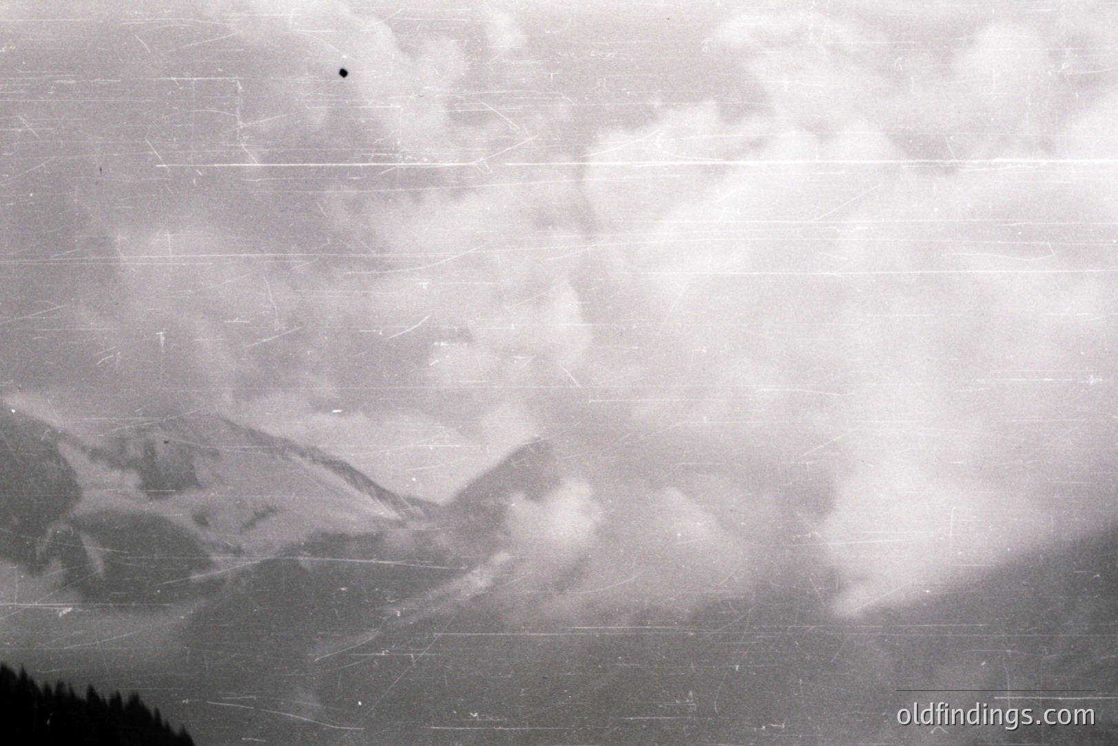 Dramatic, monochromatic view of a mountainous landscape obscured by thick, swirling clouds. Visible peaks suggest alpine terrain, potentially captured during a storm. Significant film grain and surface scratches are evident. Likely mid-20th century, amateur photography. Suitable for vintage stock or design.