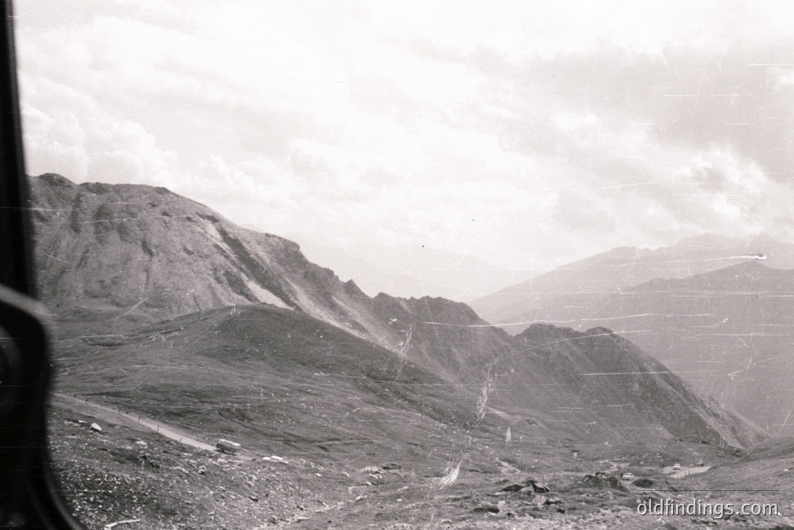 Dramatic monochrome landscape features rugged, sloped mountains receding into distance. A winding road cuts across the terrain. Likely an alpine view, possibly taken from a high vantage point. Grainy texture suggests older film stock. Likely 1960s-1970s era. Stock potential for travel or landscape design.