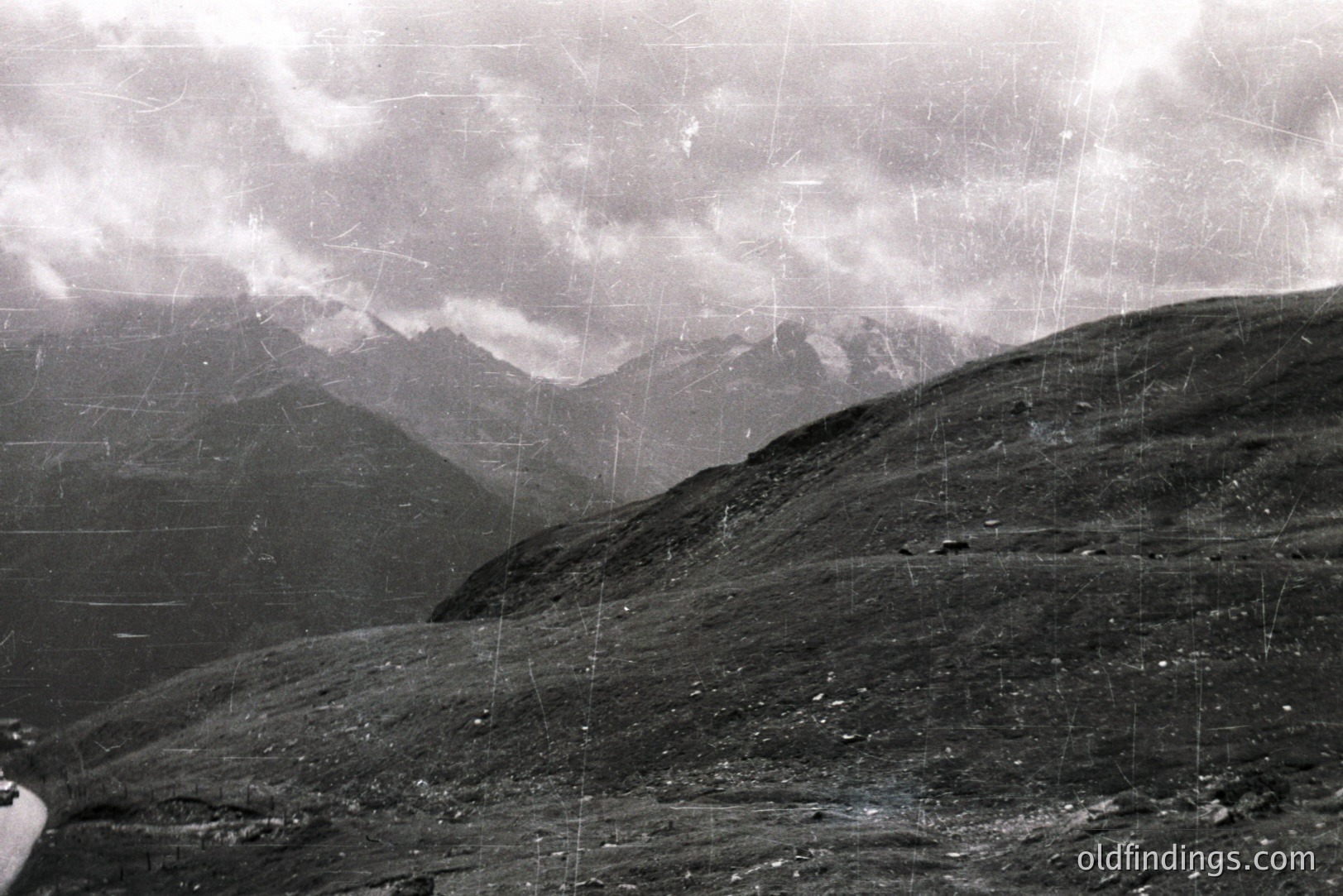 Dramatic, monochrome landscape showcasing a rugged, alpine terrain under a heavy, overcast sky. A winding road is visible in the lower-left corner, leading into the valley. The scene conveys a sense of isolation and vastness. Likely mid-20th century, possibly 1950s or 60s. Could be suitable for travel or landscape design references.