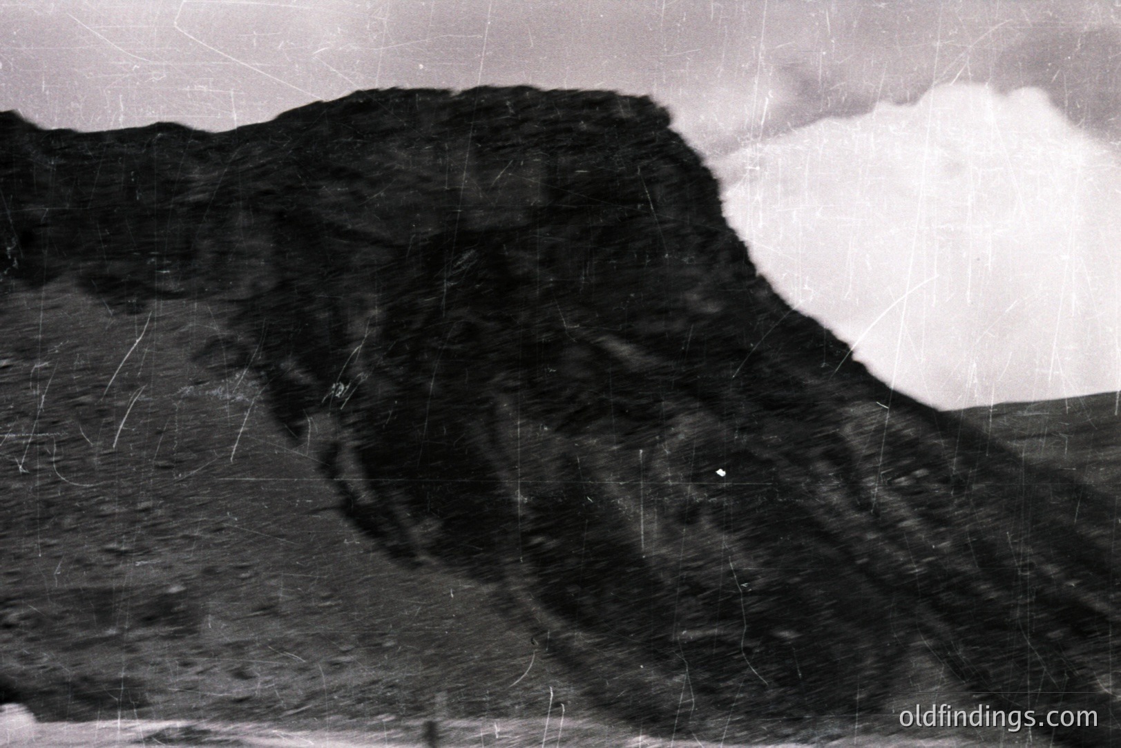 Striking black and white photo featuring a dramatic, rocky cliff face under a cloudy sky. Texture and erosion are evident on the rock surface. Likely a coastal scene, potentially taken during the mid-20th century judging by the film grain and photographic style. Appears to be a vintage print with surface scratches.
