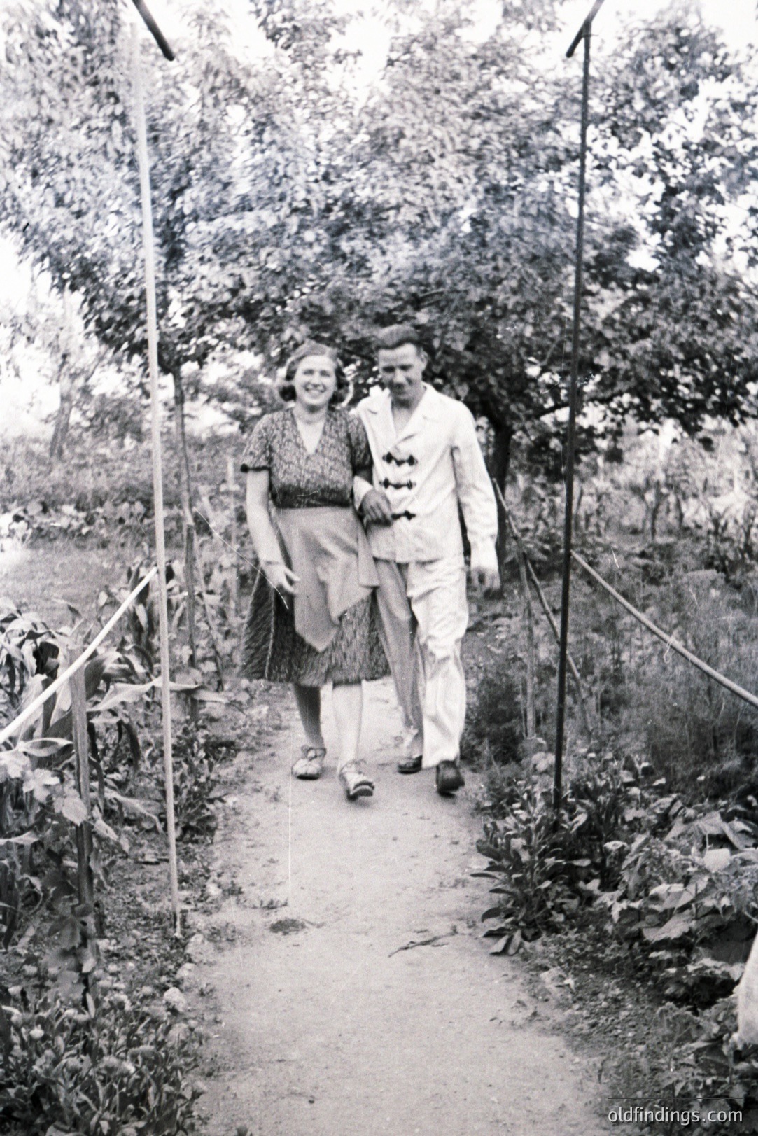 A couple strolls along a gravel path flanked by lush foliage, showcasing 1960s fashion. The woman wears a patterned dress; the man, a double-breasted white suit. A simple wire fence lines the path. Likely a resort or garden setting.