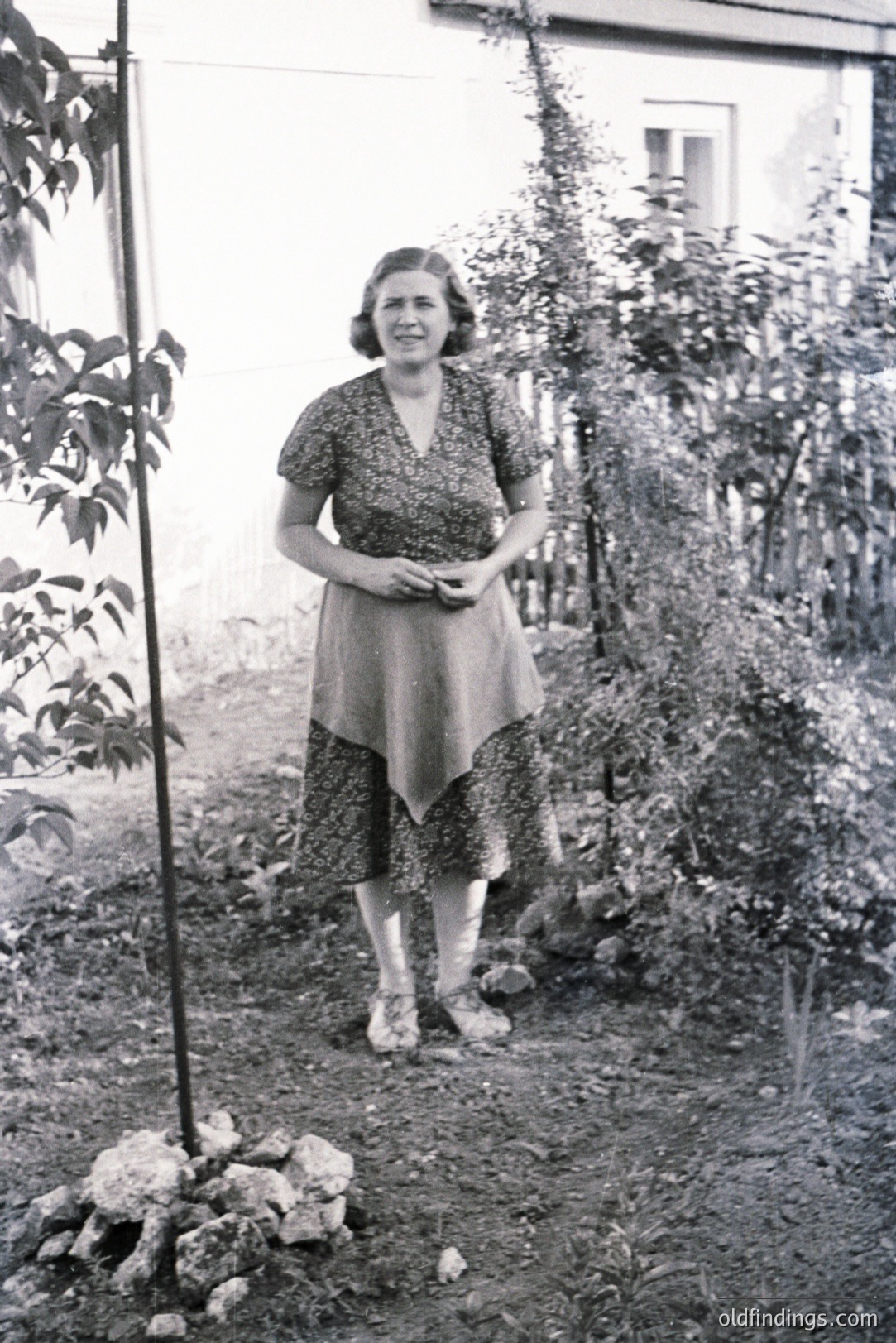 A woman stands in a garden with a stone wall and lush foliage in the background. She wears a patterned, knee-length dress with short sleeves and sandals. Likely a family portrait, possibly from the 1930s-1950s, reflecting mid-century fashion. The setting suggests a private residence.
