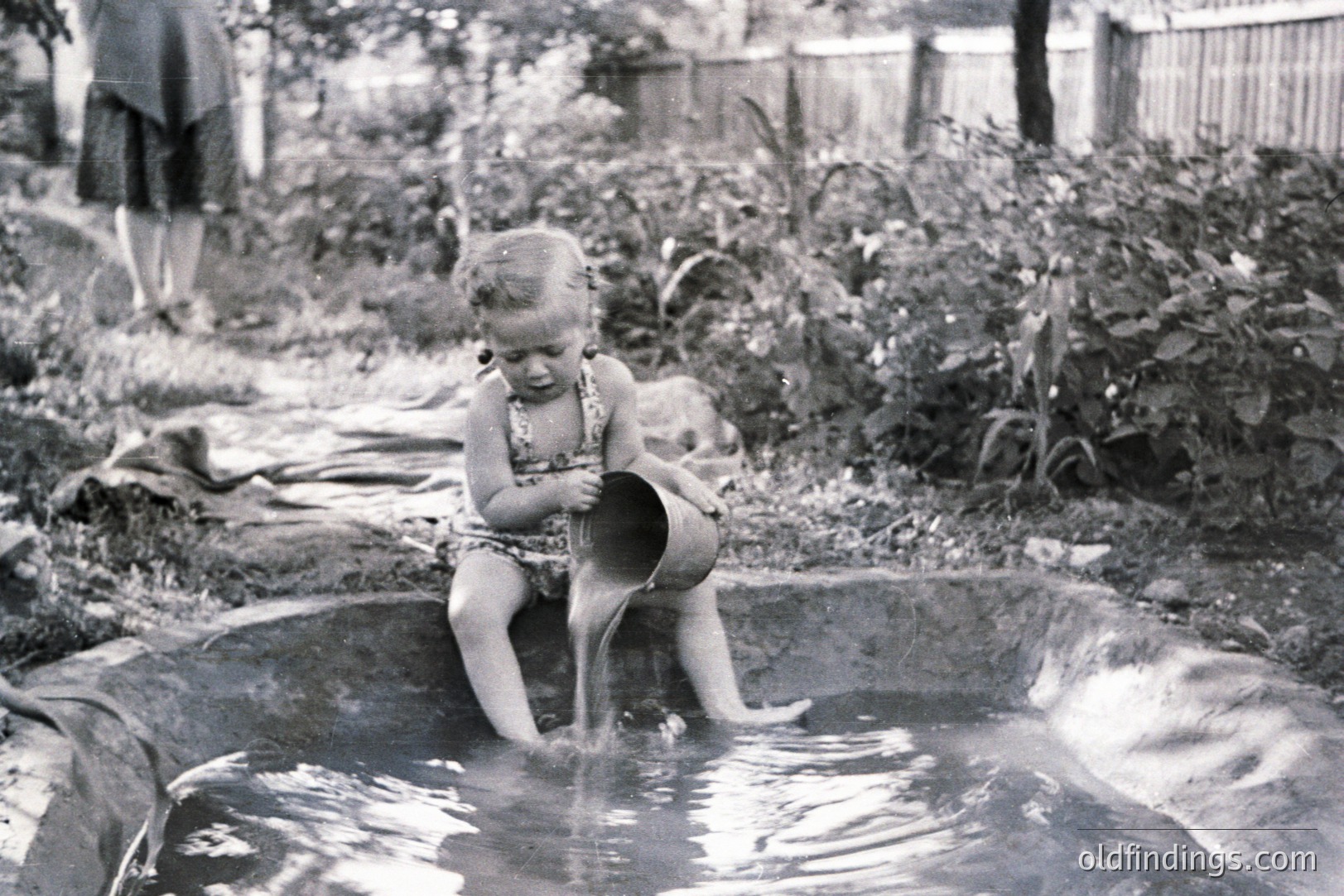 A young girl sits in a shallow, stone-lined pool, playfully pouring water from a bucket. Lush foliage fills the background, with a glimpse of a woman in a dress visible further back. Likely a candid, family snapshot, circa 1950s-1960s. Captures a moment of childhood play and a natural, backyard setting.