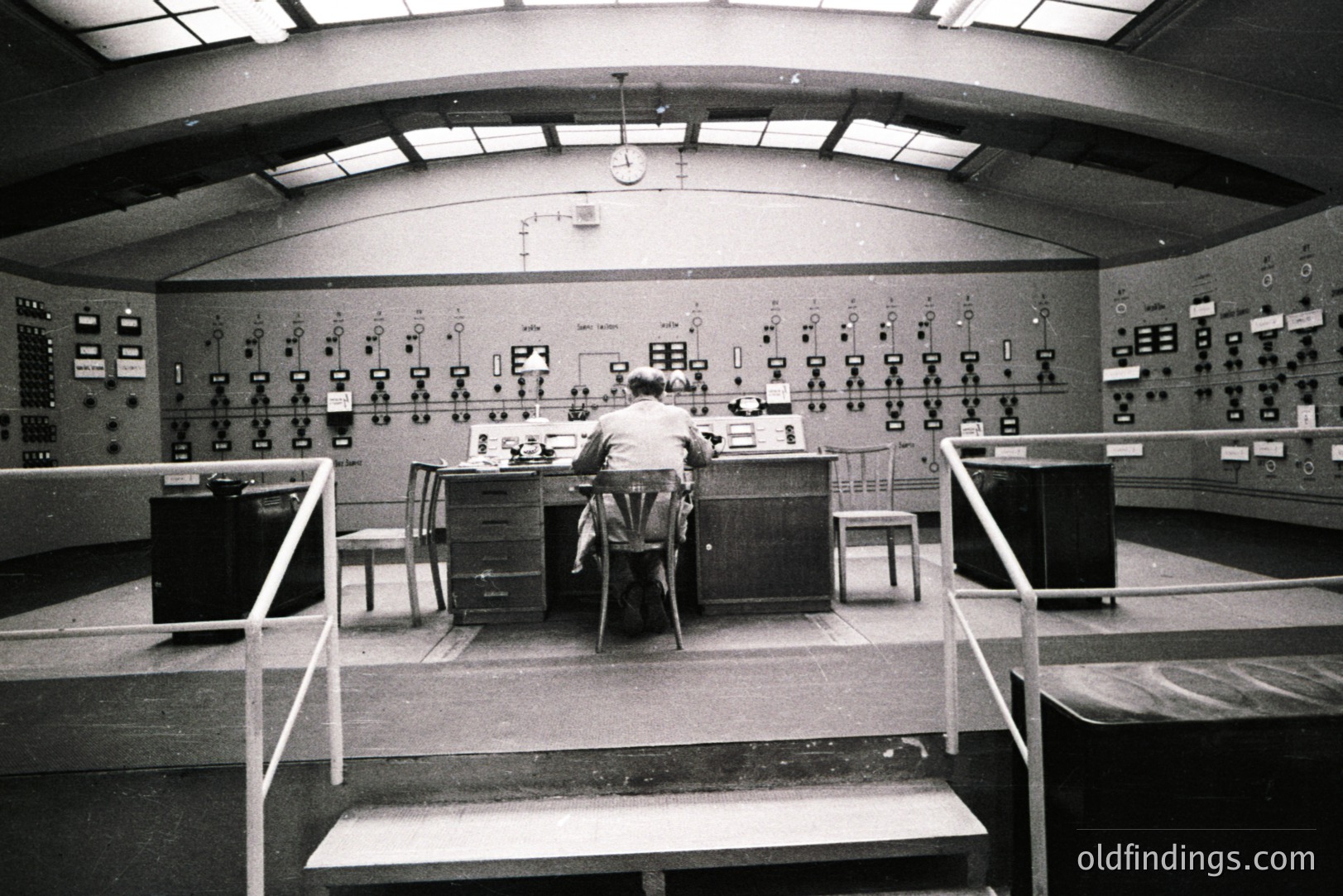 A man sits at a control panel in a large, curved room, likely an industrial or scientific facility. Numerous gauges, dials, and switches cover the wall. Concrete construction and a raised platform suggest a 1960s-70s operational environment. A clock is visible high on the wall.