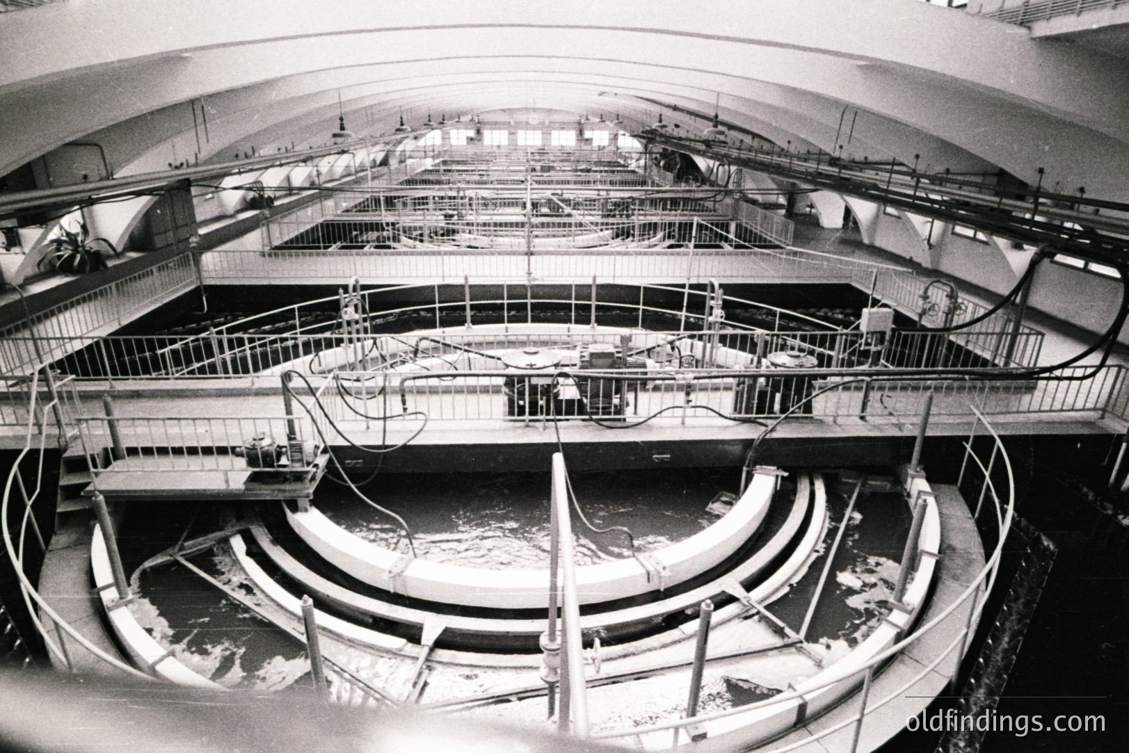 Industrial scene: circular water treatment basin with tiered platforms and railings. Visible piping and machinery suggest a filtration or processing facility. Likely 1960s-1970s architecture, possibly European. Shows engineering and infrastructure.