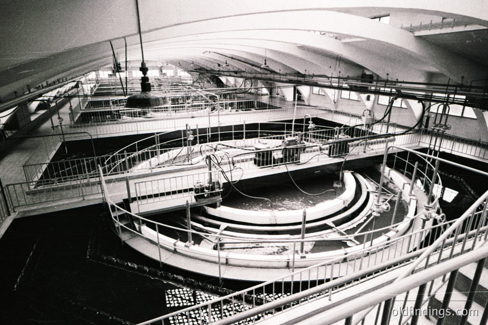 Intricate view of a large, multi-tiered pool system with a spiral central feature and raised walkways. Likely a research or aquaculture facility. The architecture features sharp angles and a high ceiling with exposed mechanical elements. Strong geometric patterns in the layout. Appears to be from the mid-20th century.