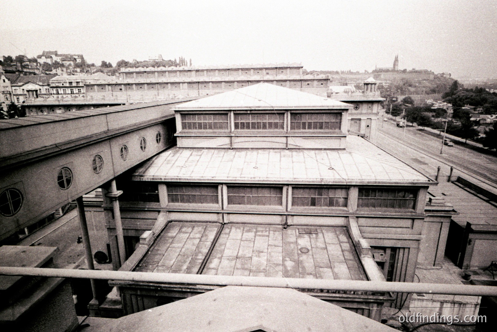A view of a large, modernist institutional building, possibly a hospital or school. Geometric architecture with flat roofs and a prominent central dome. Extensive glazing indicates natural light emphasis. Surrounding landscape shows rolling hills & distant cityscape. Appears to be circa 1930s-1950s.