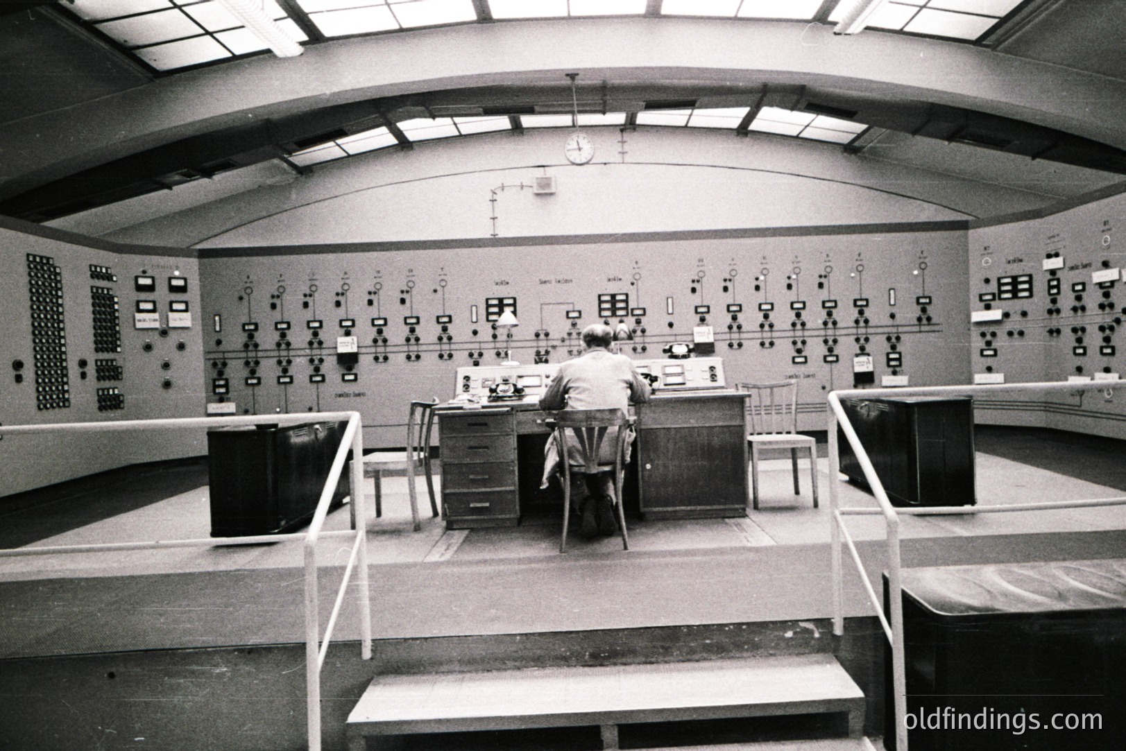 Man seated at a control panel densely populated with switches and meters within a large, high-ceilinged room. Likely a telecommunications or industrial control center. The architectural style suggests a mid-century design (1960s-1970s). Sparse furnishings, utilitarian aesthetic. Industrial function over form.