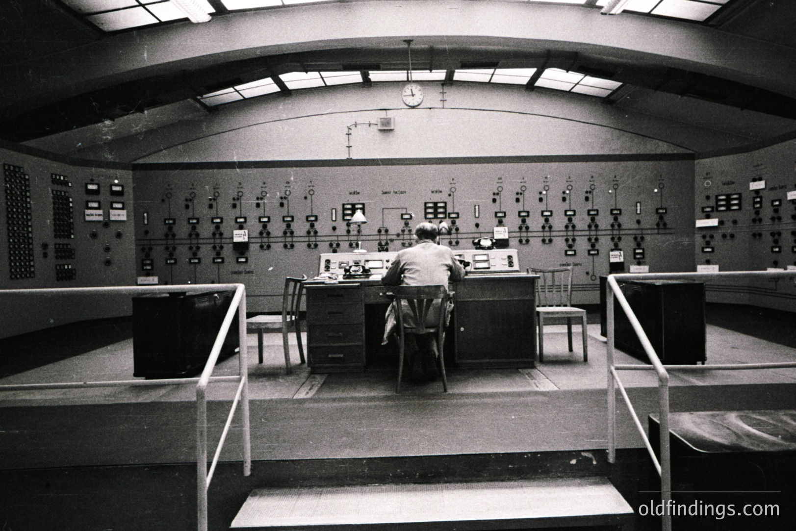 A technician sits at a control console within a large, industrial room. Numerous panel meters and displays cover the walls, suggesting a power plant or communications facility. Elevated walkway and ribbed ceiling indicate a modernist, functional design. Likely 1960s-1970s industrial aesthetic.