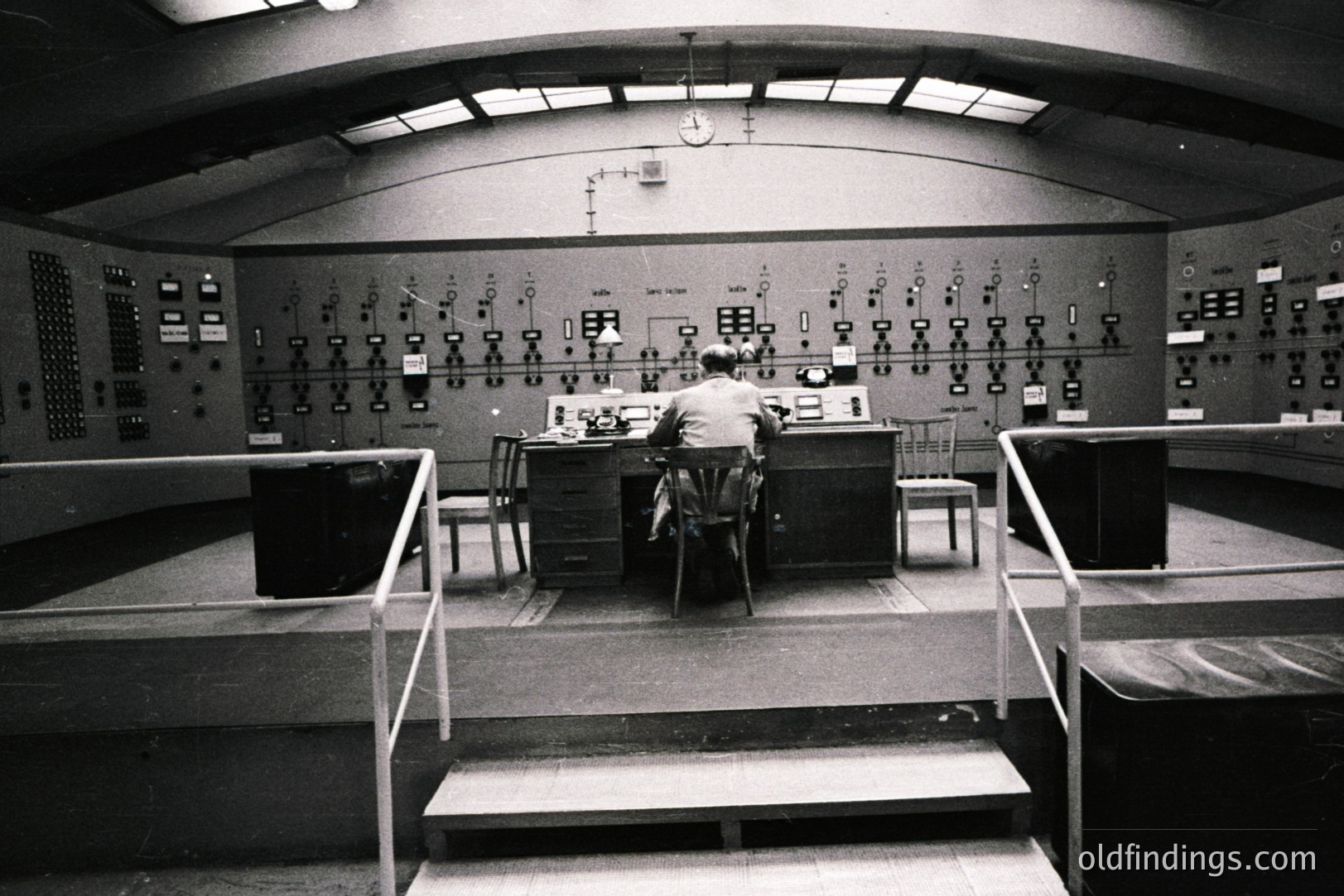 A technician sits at a control panel within a stark, elevated room. Numerous gauges, switches, and indicator lights cover the large wall, suggesting a technological or industrial setting. The elevated platform, handrails, and arched ceiling indicate a specialized facility. Likely 1960s-1970s.