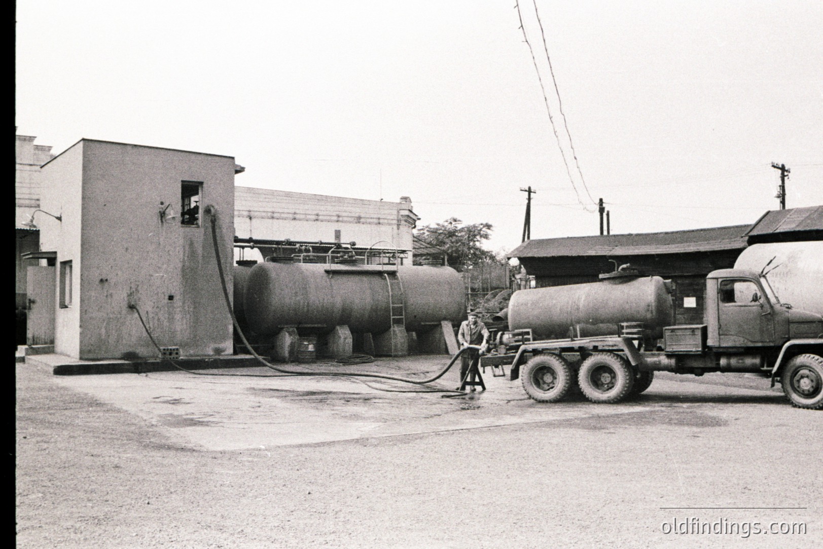 Industrial scene: Large fuel storage tanks dominate the view, with a tanker truck parked beside them, attended by a worker. Simple, functional architecture suggests an oil depot or similar facility. Likely mid-20th century, potentially post-WWII industrial expansion. Appears to be a working site, capturing routine operations.