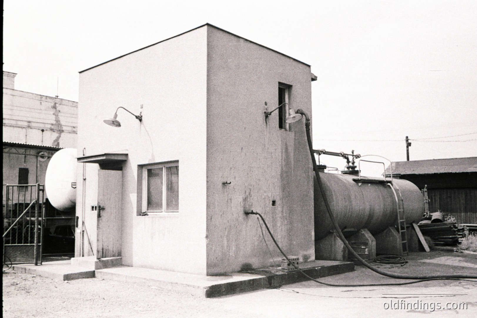 Industrial complex with a monolithic concrete structure, featuring a large propane tank and connected piping system. The building's stark architectural style suggests post-war utilitarian design. Likely a storage or processing facility. Possible mid-20th century, Eastern European location.
