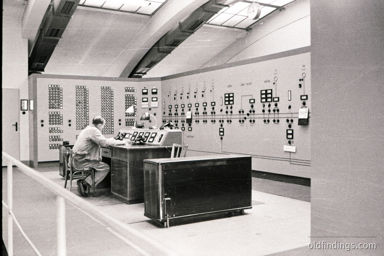 Control room interior, likely an industrial facility. Two men seated at a large console monitoring complex control panels filled with numerous switches & displays. The room exhibits a minimalist, functional design with a curved ceiling. Architecture suggests 1960s or 70s industrial aesthetic. Potential stock photo reference.