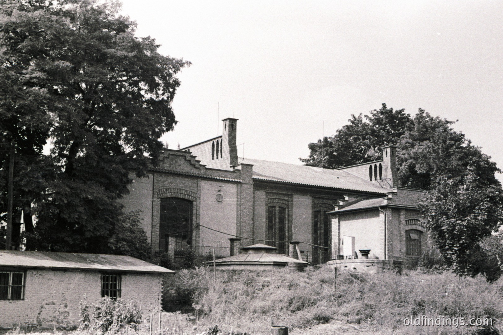 Elegantly designed brick villa with arched windows, chimneys, and a small turret. Visible evidence of architectural detail and surrounding overgrown vegetation suggest a stately estate. Likely a European location, possibly early to mid-20th century. The building's style points to potential design influence.