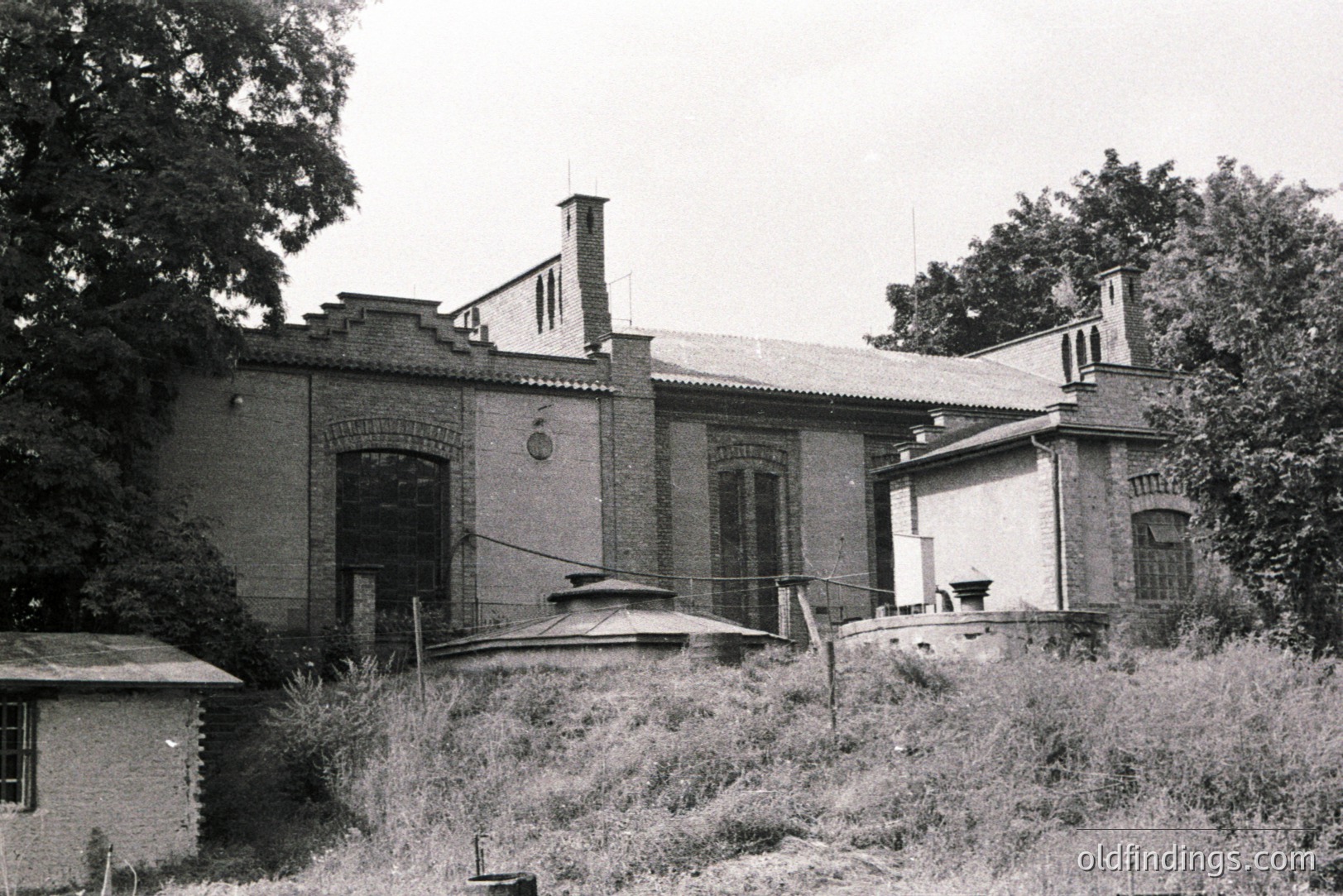 A brick building with elaborate detailing, including a round turret and decorative chimneys, sits amidst overgrown vegetation. Likely a former estate or industrial building, the architecture showcases a mix of styles. Possible location: Eastern Europe, judging by the brickwork & detailing.
