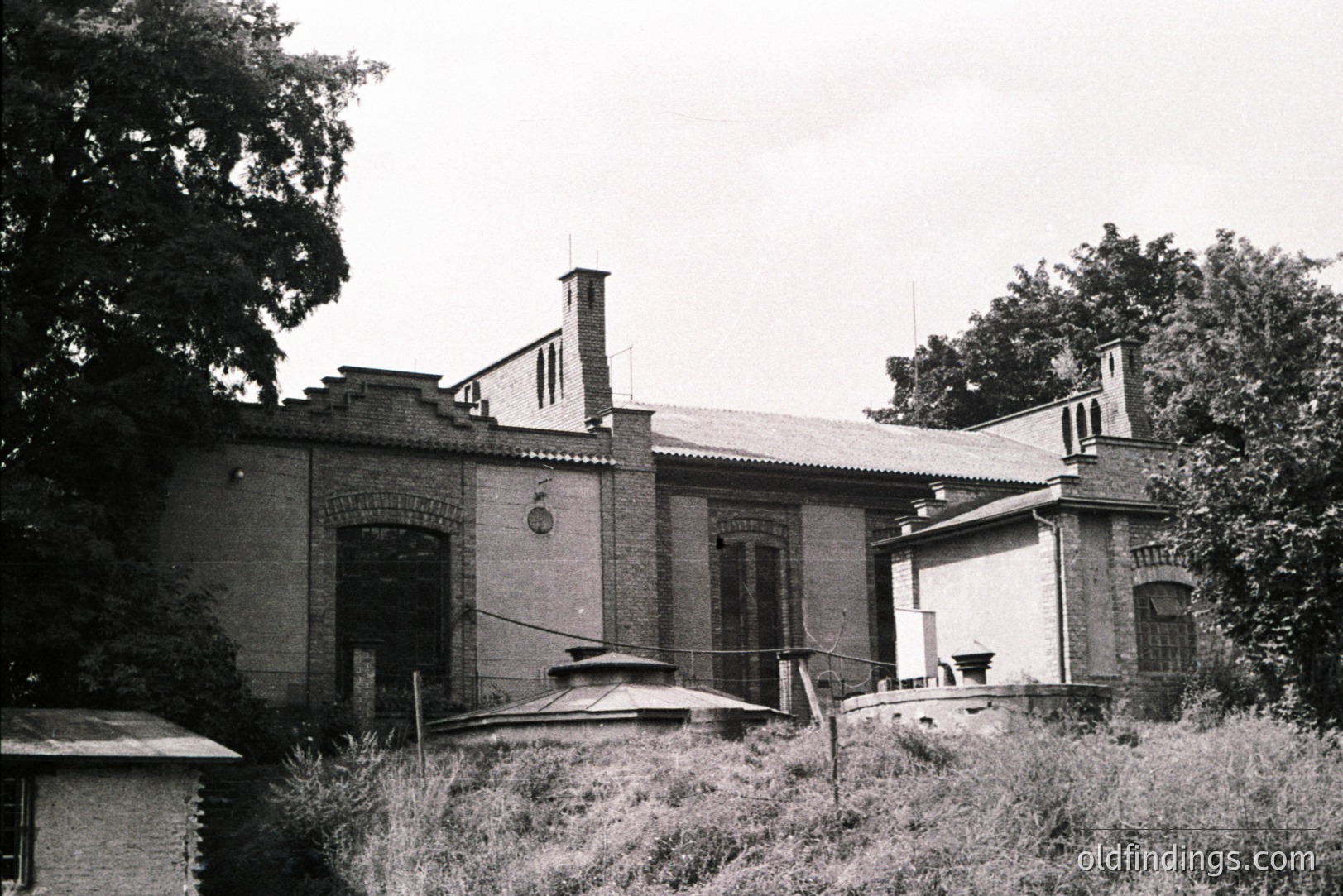 A two-story brick building with arched windows and decorative stonework, set within overgrown vegetation. Note the prominent chimney and the detailed cornice. Likely a villa or estate building, possibly early 20th century. Reflects regional architectural styles. Potential for stock use in design/historical settings.