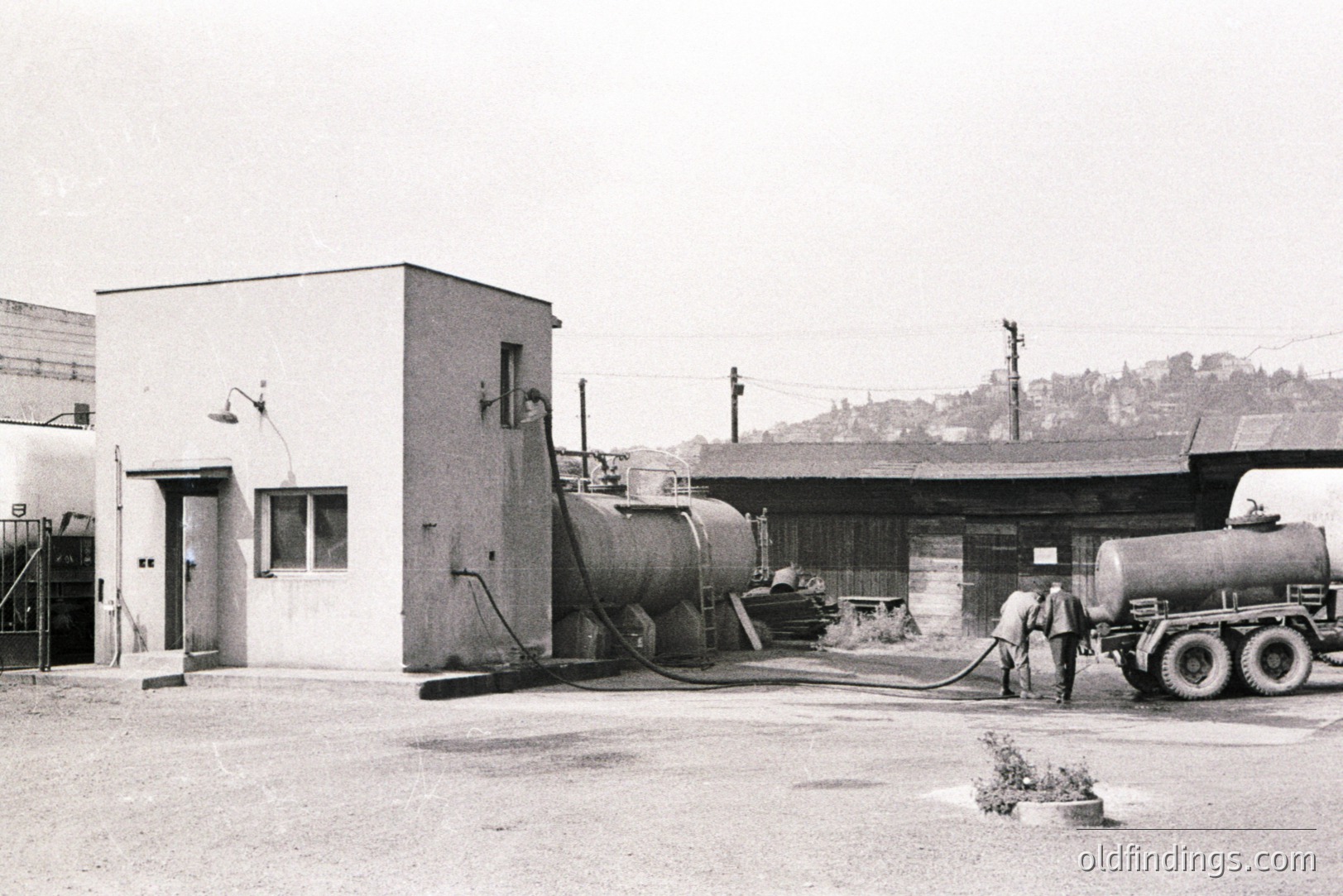 Industrial landscape featuring large storage tanks and a utilitarian building with a prominent chimney. Appears to be a processing or storage facility. Background shows a hillside town. Likely 1950s-1970s, potentially Eastern European. Suitable for design or historical context.