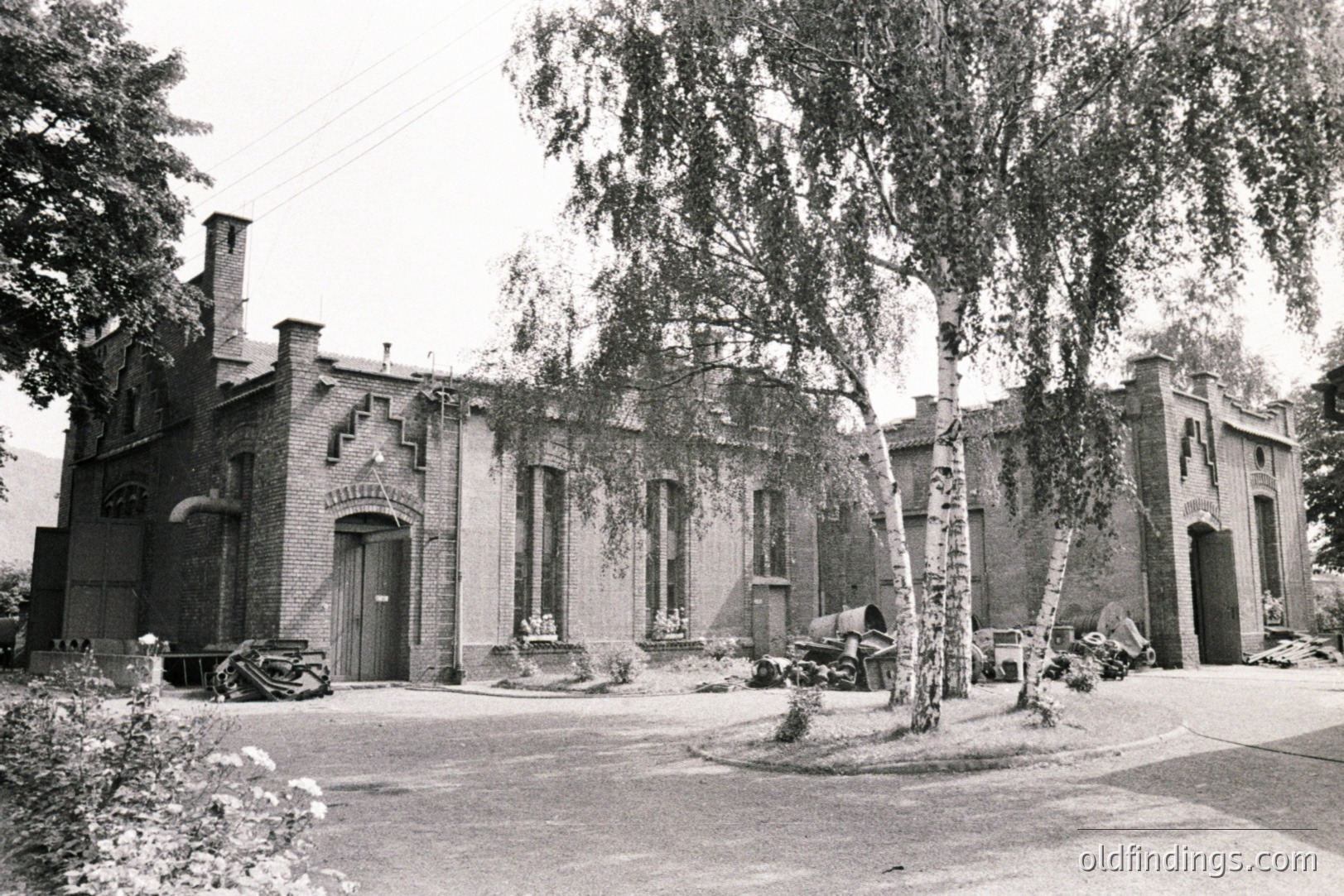 A view of a brick industrial building with distinctive crenellations and arched doorways. Adjacent workshops are visible, with assorted equipment scattered outside. The structure appears to be situated on a paved area, with mature birch trees framing the scene. Likely a factory or storage facility, c. 1970s.