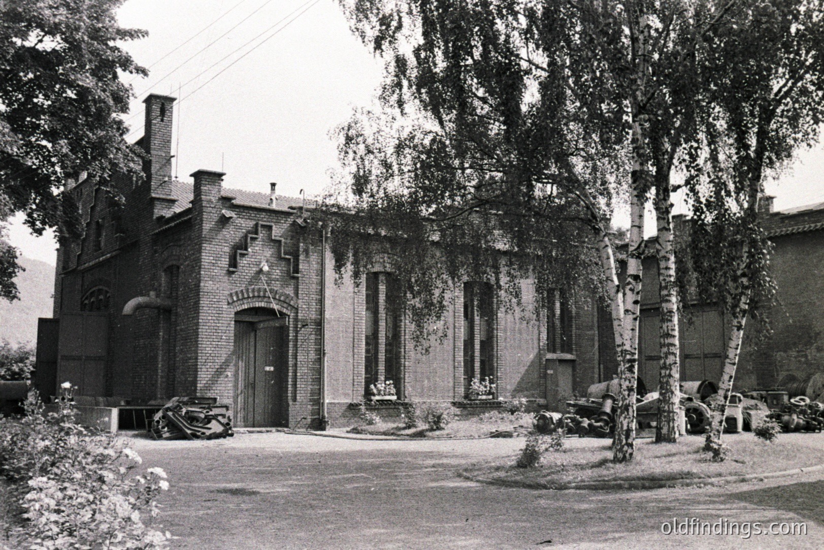 Detailed image of a brick building with elaborate gabled entrance and window detailing. Appears to be a factory or workshop, with stacked materials visible near exterior walls. Likely mid-20th century, possibly 1950s. Courtyard setting with birch trees. Commercial/industrial architectural reference.