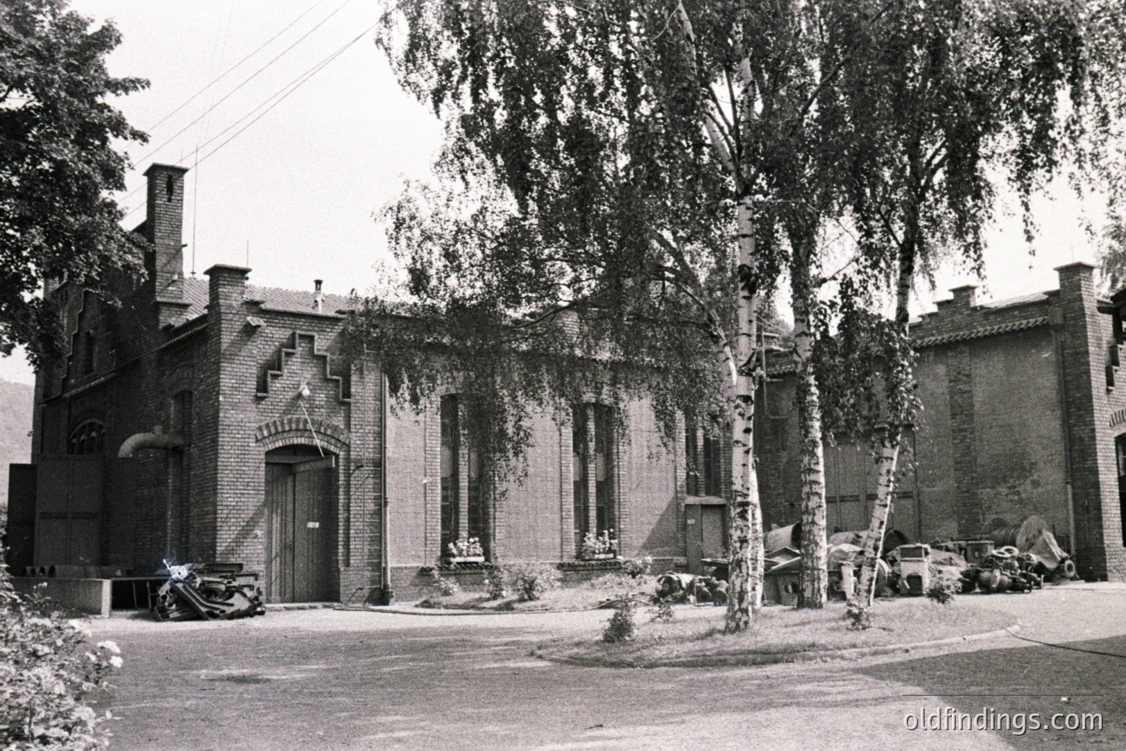 A brick building with arched entry and decorative detailing faces a tree-lined street. A vintage car is parked nearby. Likely a local government or civic structure, circa 1960s-70s. Architectural style blends neo-gothic and functionalist elements. Shows a small, manicured front lawn.