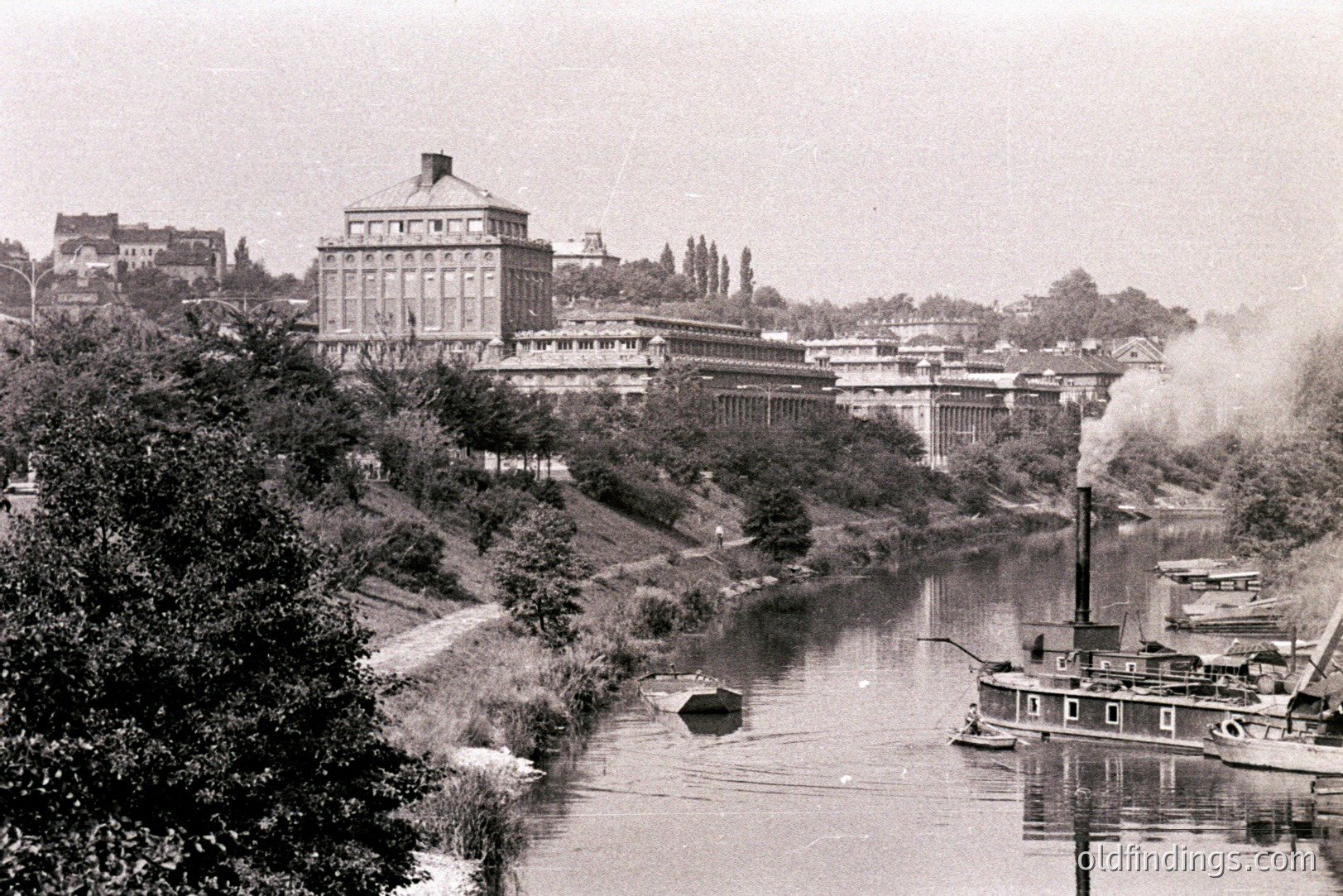 A lengthy, multi-story building with a central tower dominates the view, set within a landscape of trees and foliage. A river runs alongside, featuring a steamboat emitting steam. Likely a European location, possibly late 19th/early 20th century, suggesting industrial/resort significance.