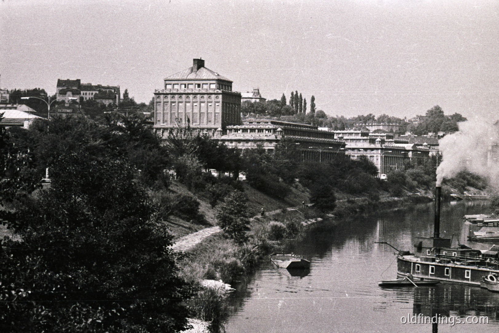 A large industrial building with a central cupola dominates a riverside scene. A steamboat emits steam on the waterway, suggesting industrial activity. Landscape with vegetation on a slope rises to the building. Appears to be late 19th/early 20th century.