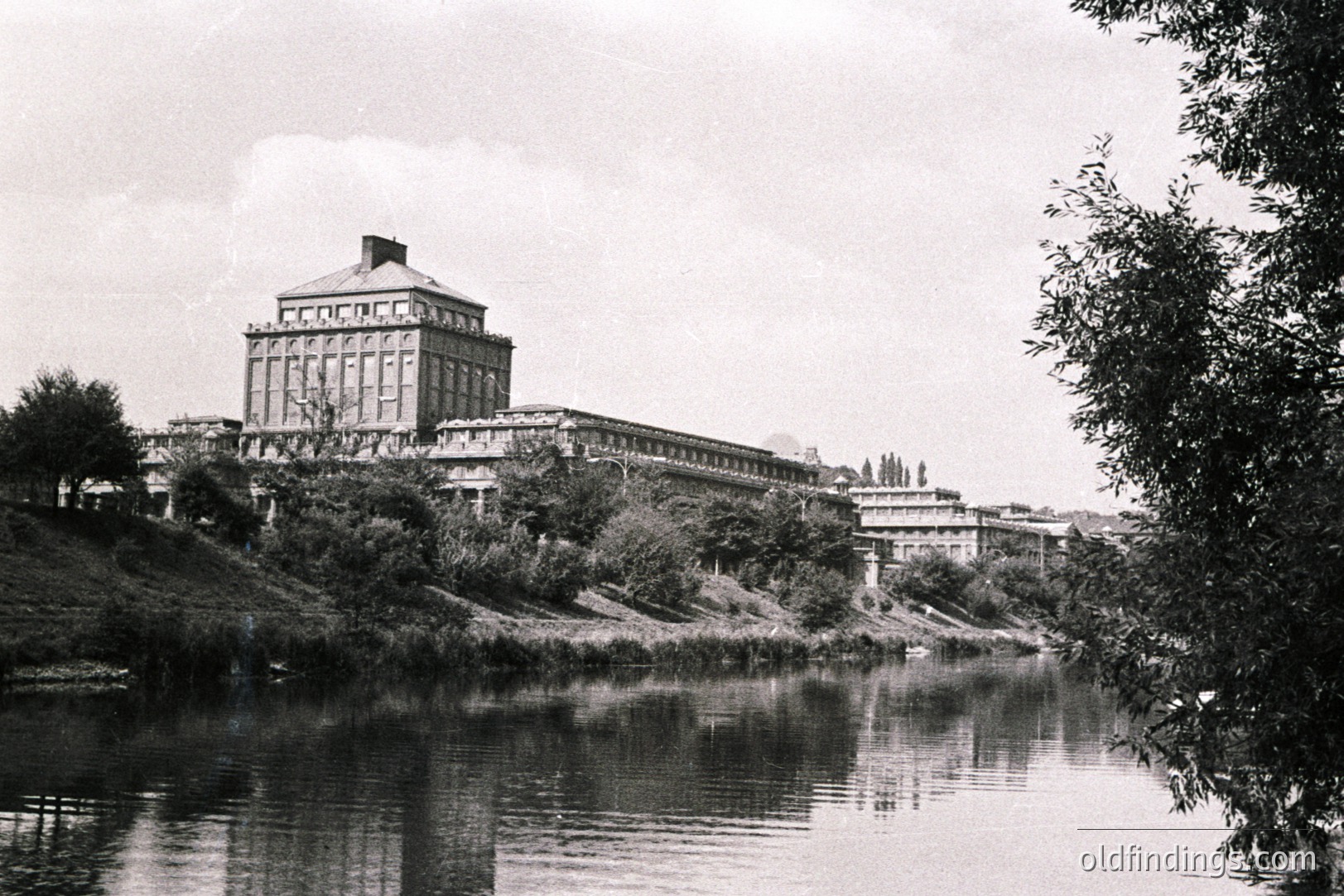 Architectural view of a grand, multi-story building with a prominent central tower. Situated on an elevated riverbank, it features symmetrical design with numerous windows and a formal landscaping. Likely a governmental or institutional building from the early-mid 20th century.