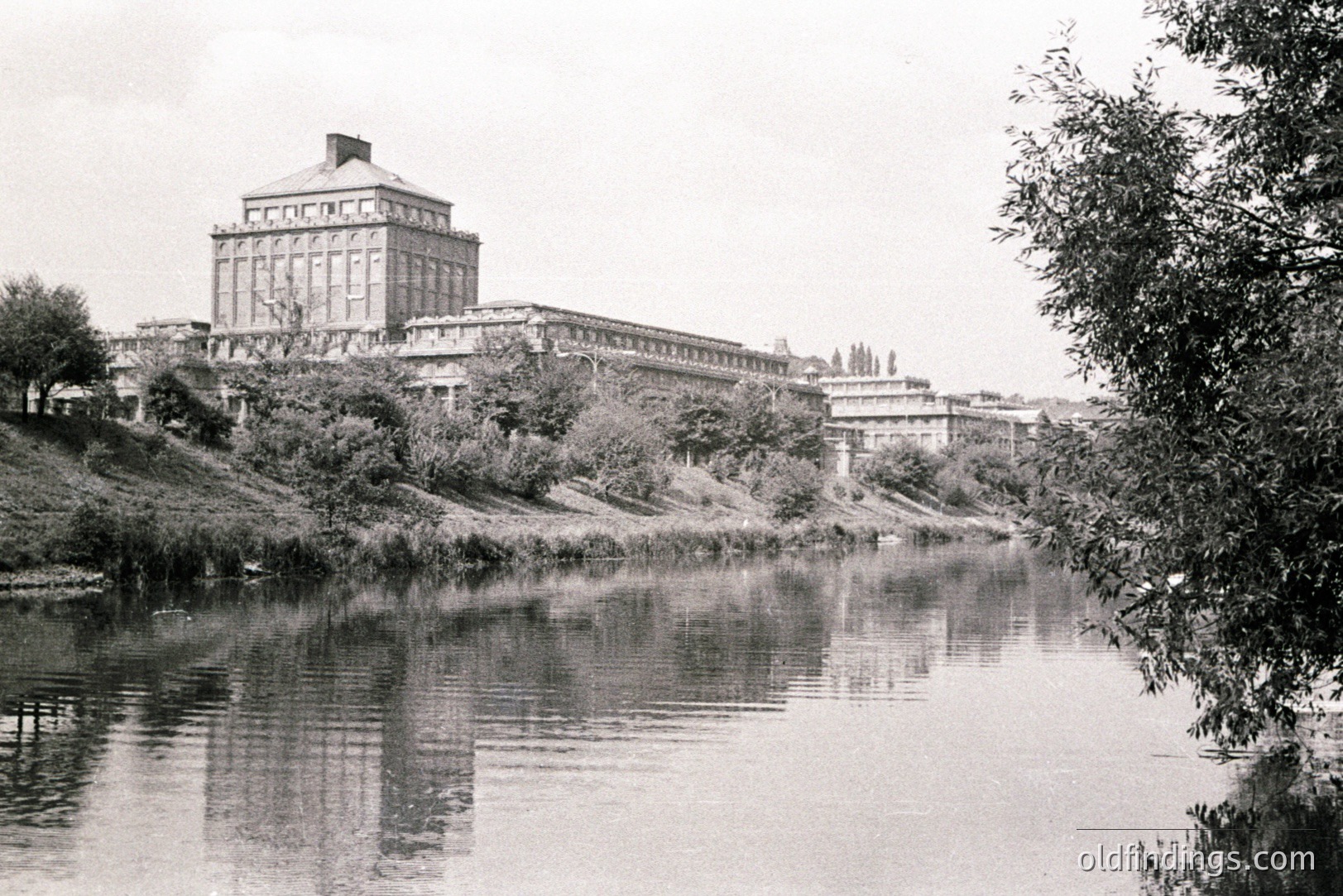 A monumental, classically-inspired building dominates the view, reflected in a still river. Extensive stonework, symmetrical facade, and a central cupola are prominent. Likely a governmental or institutional structure, possibly from the mid-20th century. Lush riverbank vegetation adds context.