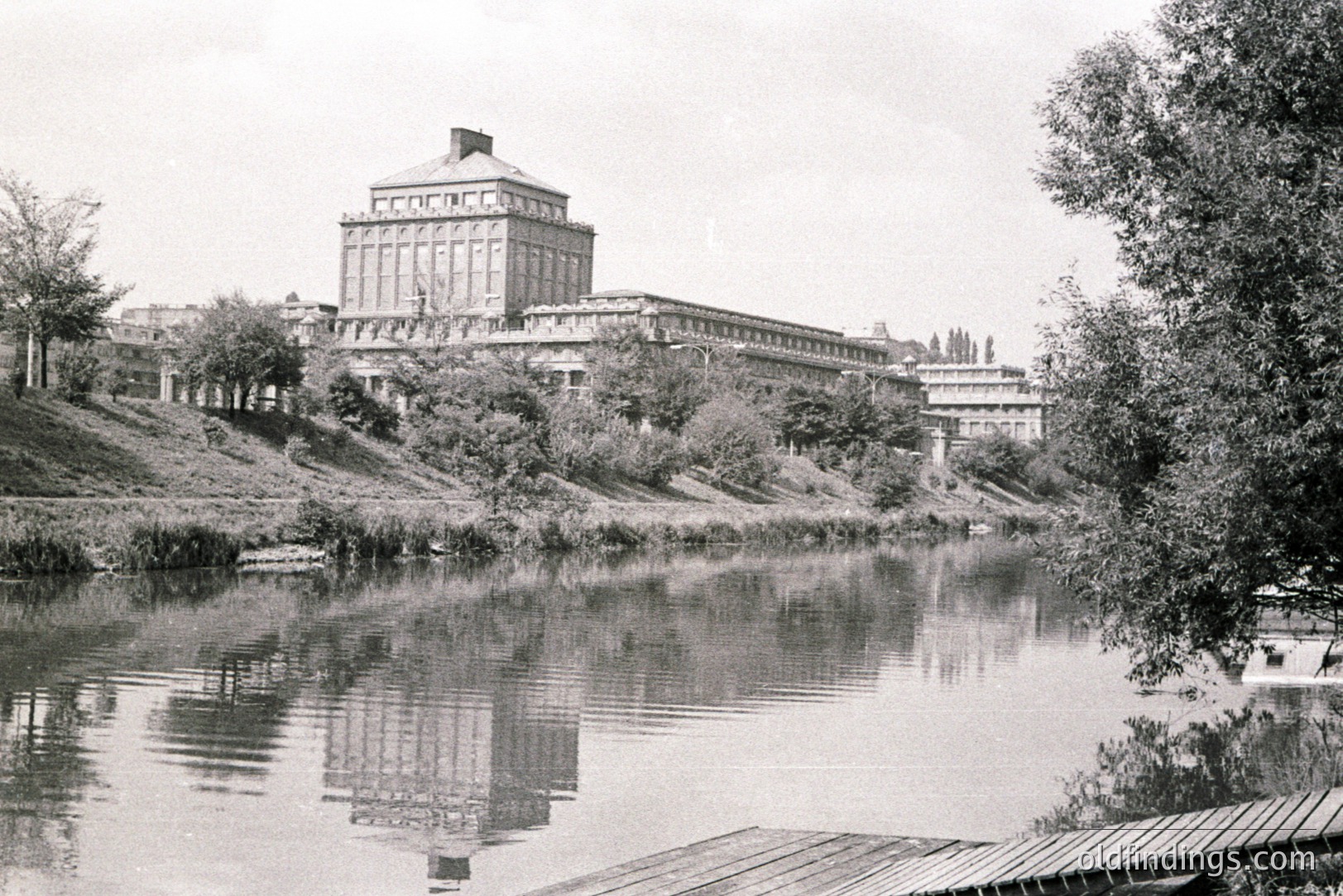 A grand, symmetrical building with a central tower dominates the view, reflected in the still waters of a lake. Architectural style suggests late 1960s-70s Eastern European design. Lush vegetation frames the composition. Likely a public institution or university.