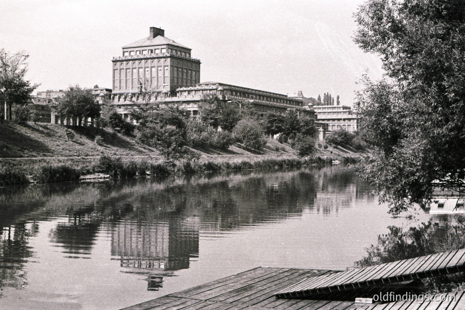 A grand, neoclassical building with a central cupola dominates this landscape, reflected in a calm body of water. Likely a public building or university, it sits elevated above the waterline, surrounded by foliage. The aged photograph suggests a mid-20th century aesthetic. This image could be valuable for architectural studies or design inspiration.